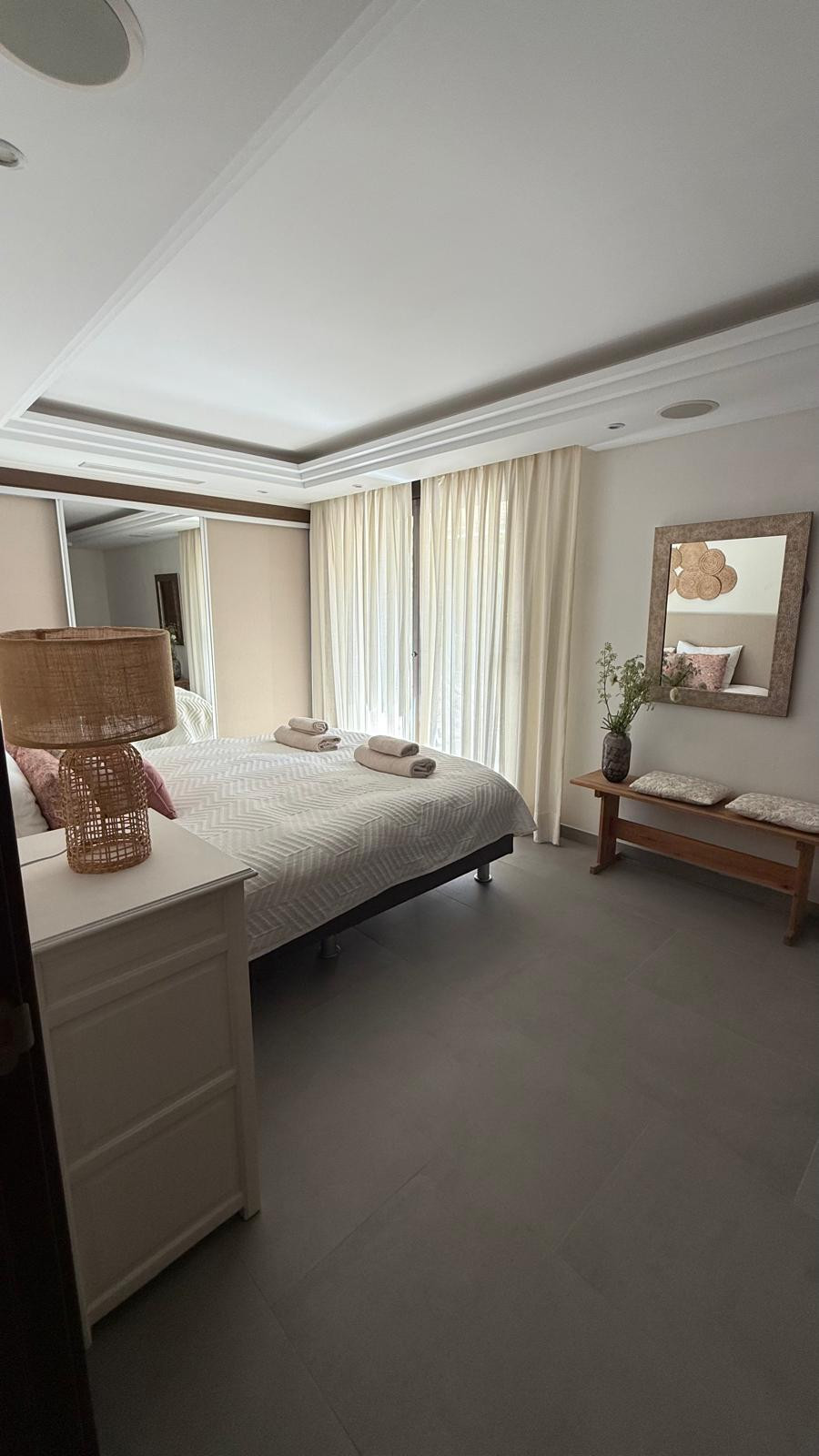 Bright bedroom with king bed, large window dressed in light curtains, and a decorative mirror.
