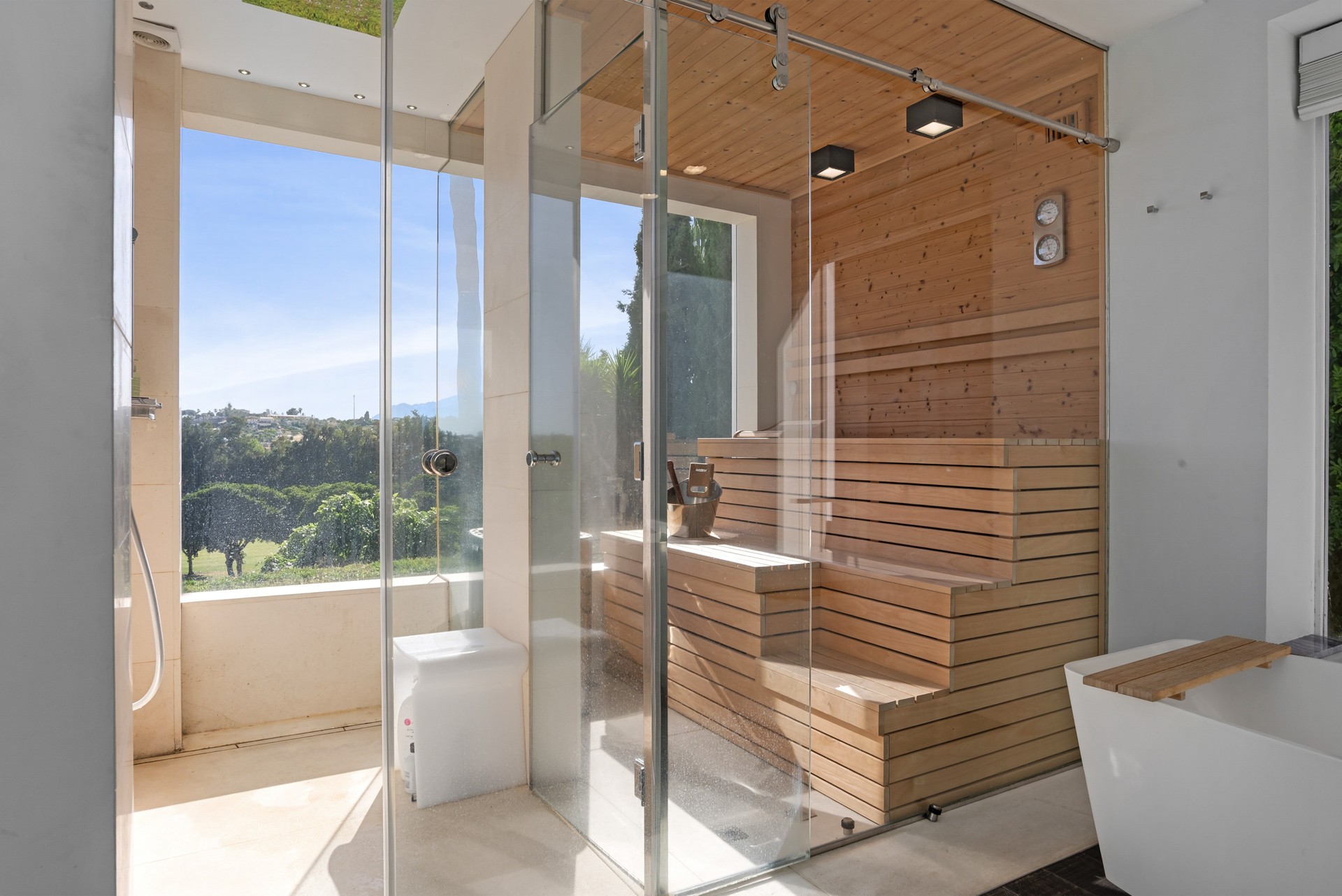 Spacious bathroom with shower, wooden sauna, and large glass windows offering views of greenery.