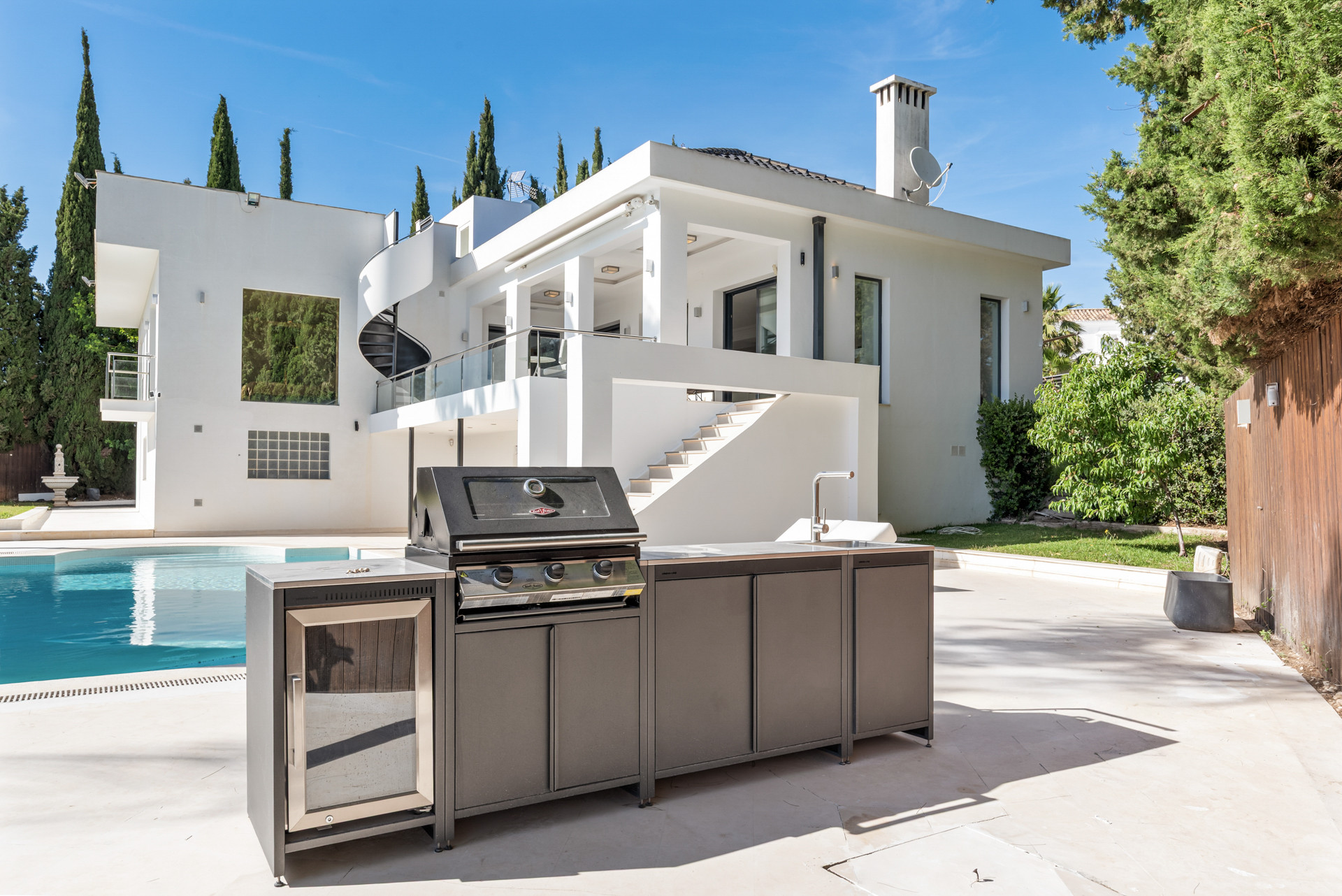 Outdoor kitchen next to the pool with a grill and sink, showcasing part of the house and surrounding greenery.