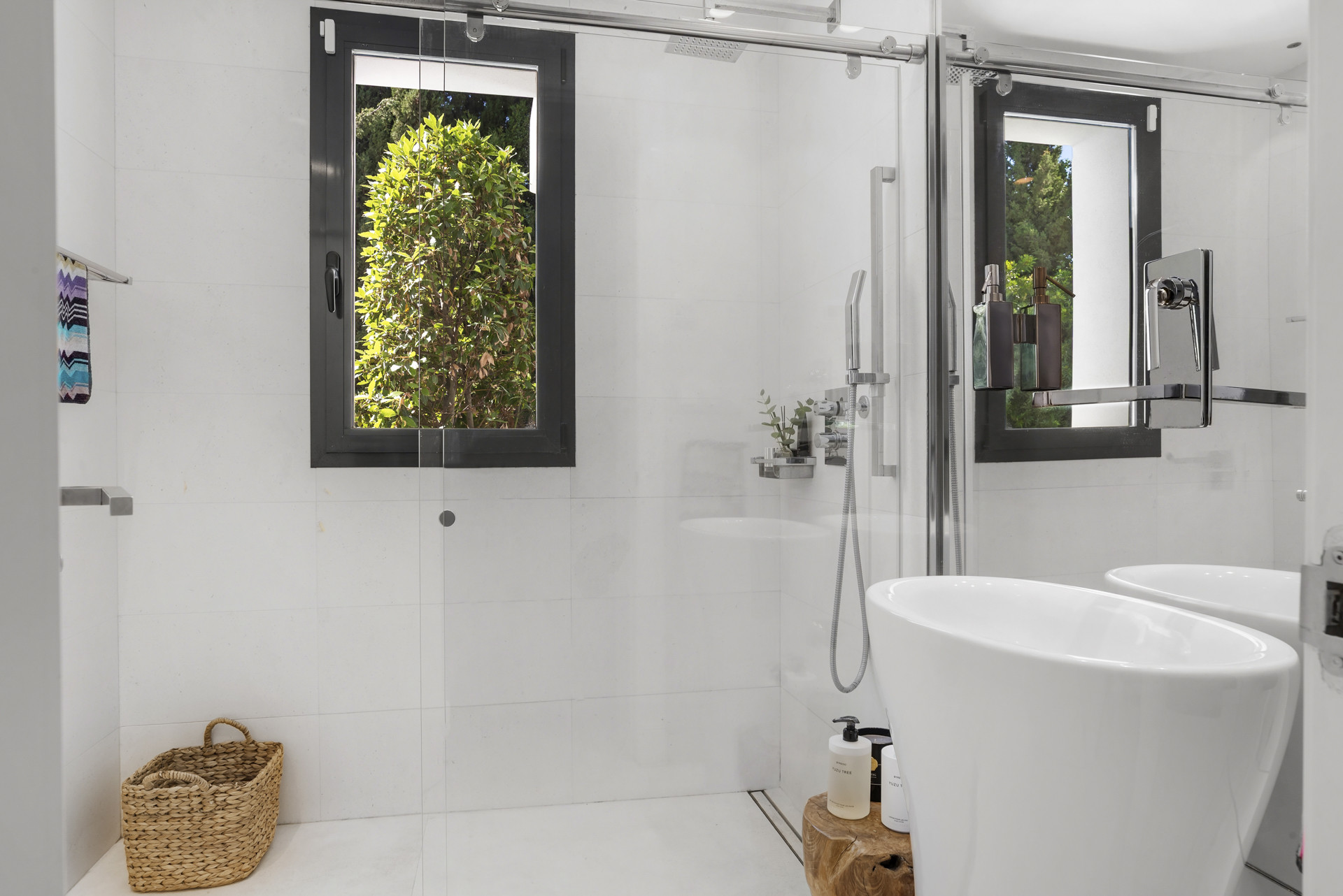 Modern bathroom with glass shower, dual sinks, and a view of greenery through the window.