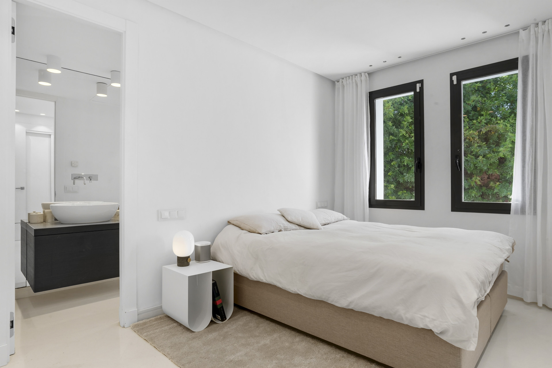 Bright bedroom with two windows and a modern bed, open bathroom area visible in the background.