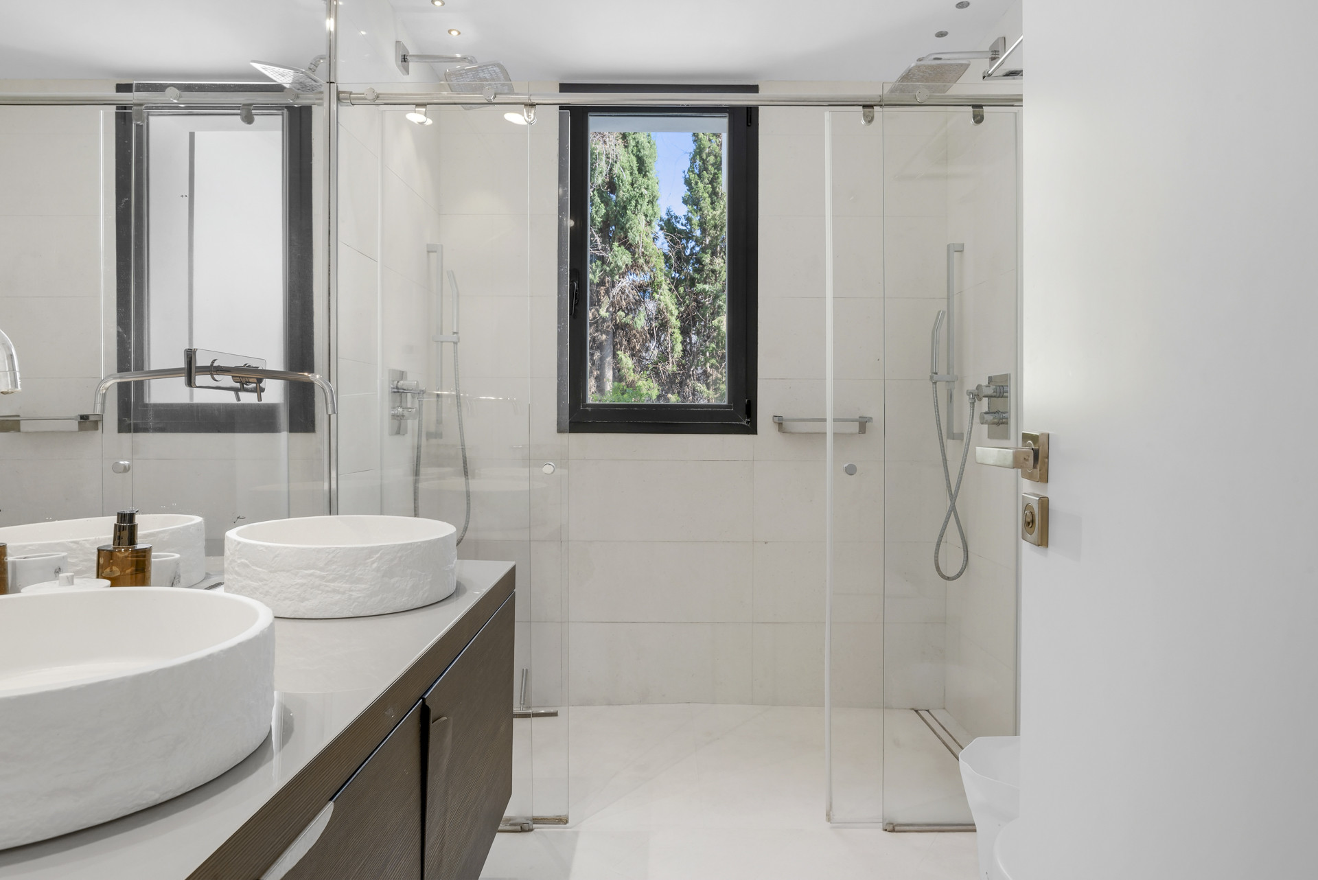 Bright bathroom equipped with two circular sinks, glass shower, and window with a view of trees.