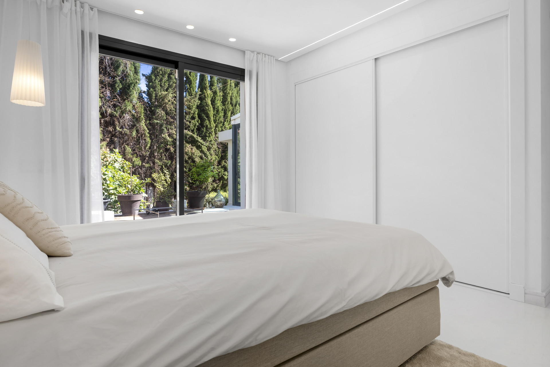 Bedroom with large windows overlooking greenery and soft lighting fixtures.