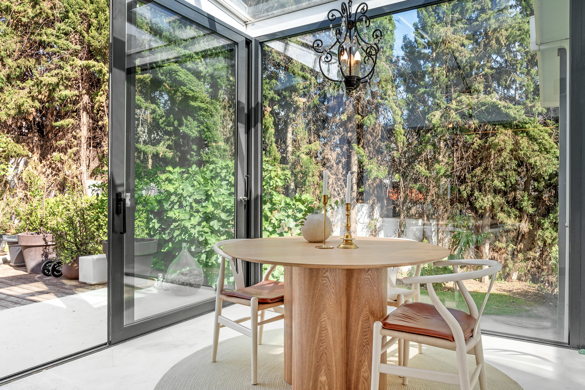 Bright dining area with a round table and chandelier, surrounded by large glass doors and greenery.