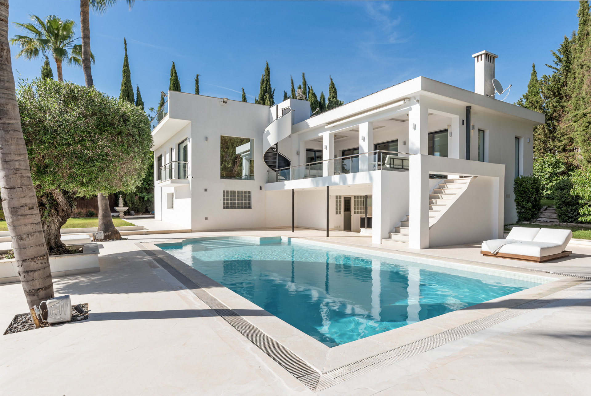 Exterior view of the house with a clear swimming pool, terrace, and lush landscaping under a blue sky.