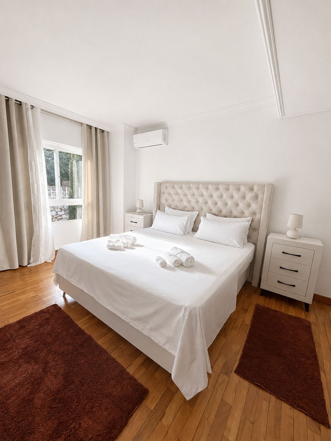 Cozy room with a plush bed and dark rugs next to natural light from a window.