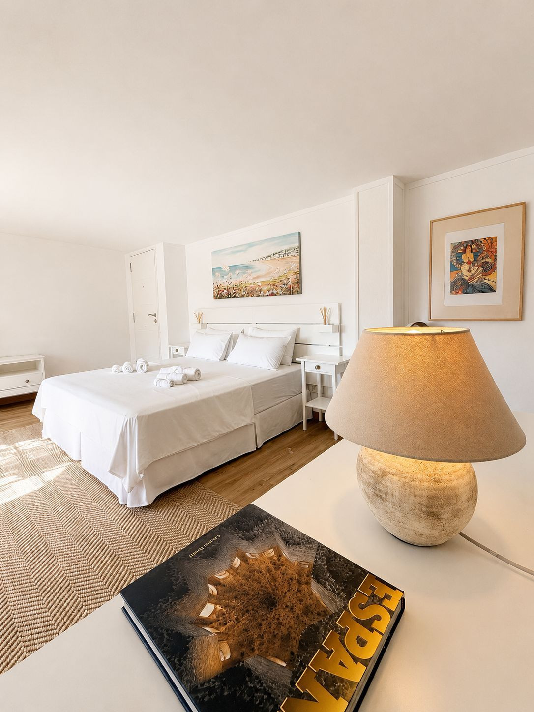 Spacious bedroom with a light decor, artwork, and a bedside lamp on a white table.