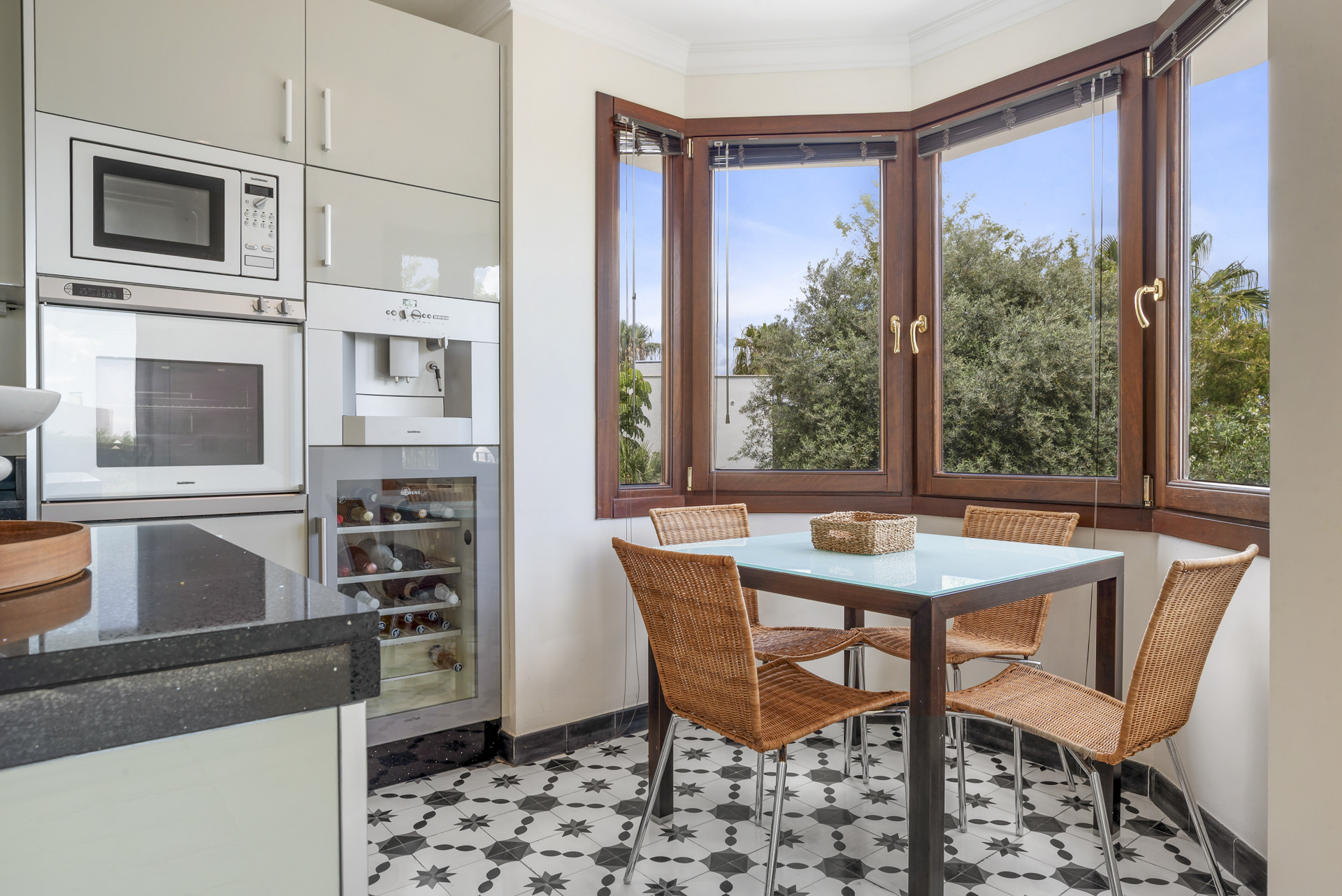 Cozy dining nook with a glass table and woven chairs, surrounded by large windows.