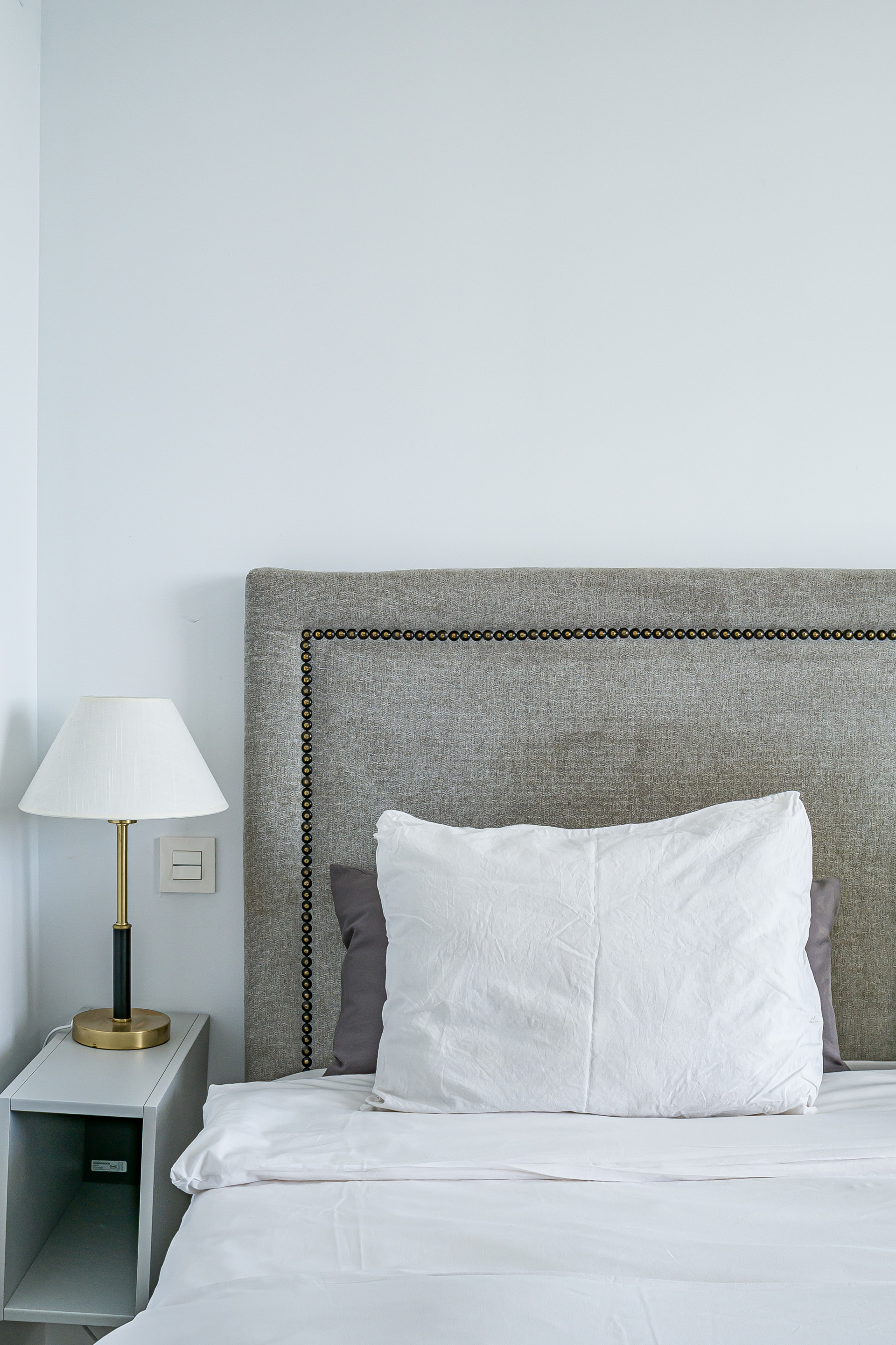 Close up of a bed with a gray upholstered headboard, white pillow, and a bedside lamp.