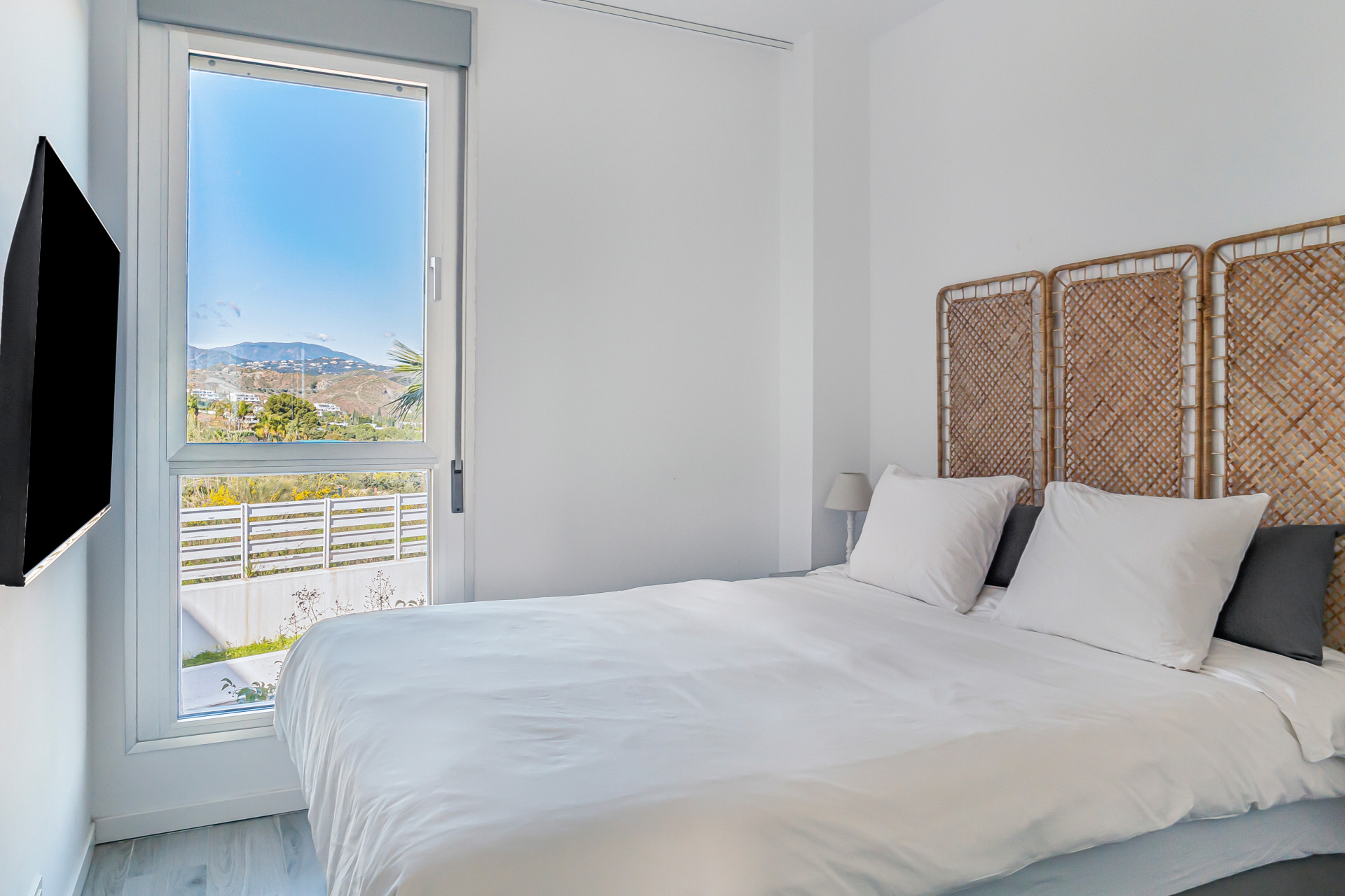 Bedroom with white bedding, wicker headboard, mounted TV, and window view of hills.