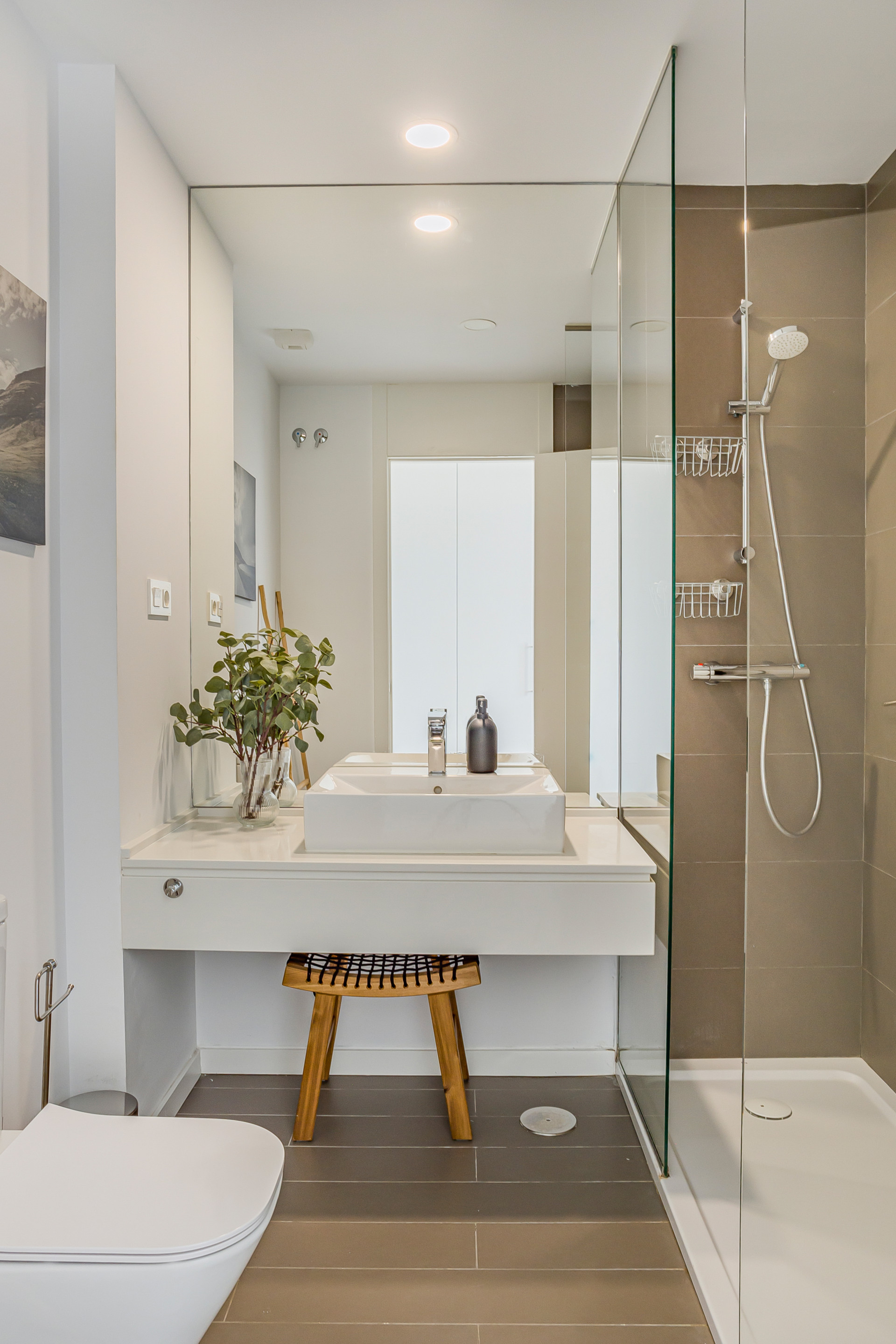 Bathroom with a white vanity sink, large mirror, wooden stool, toilet, and glass shower.