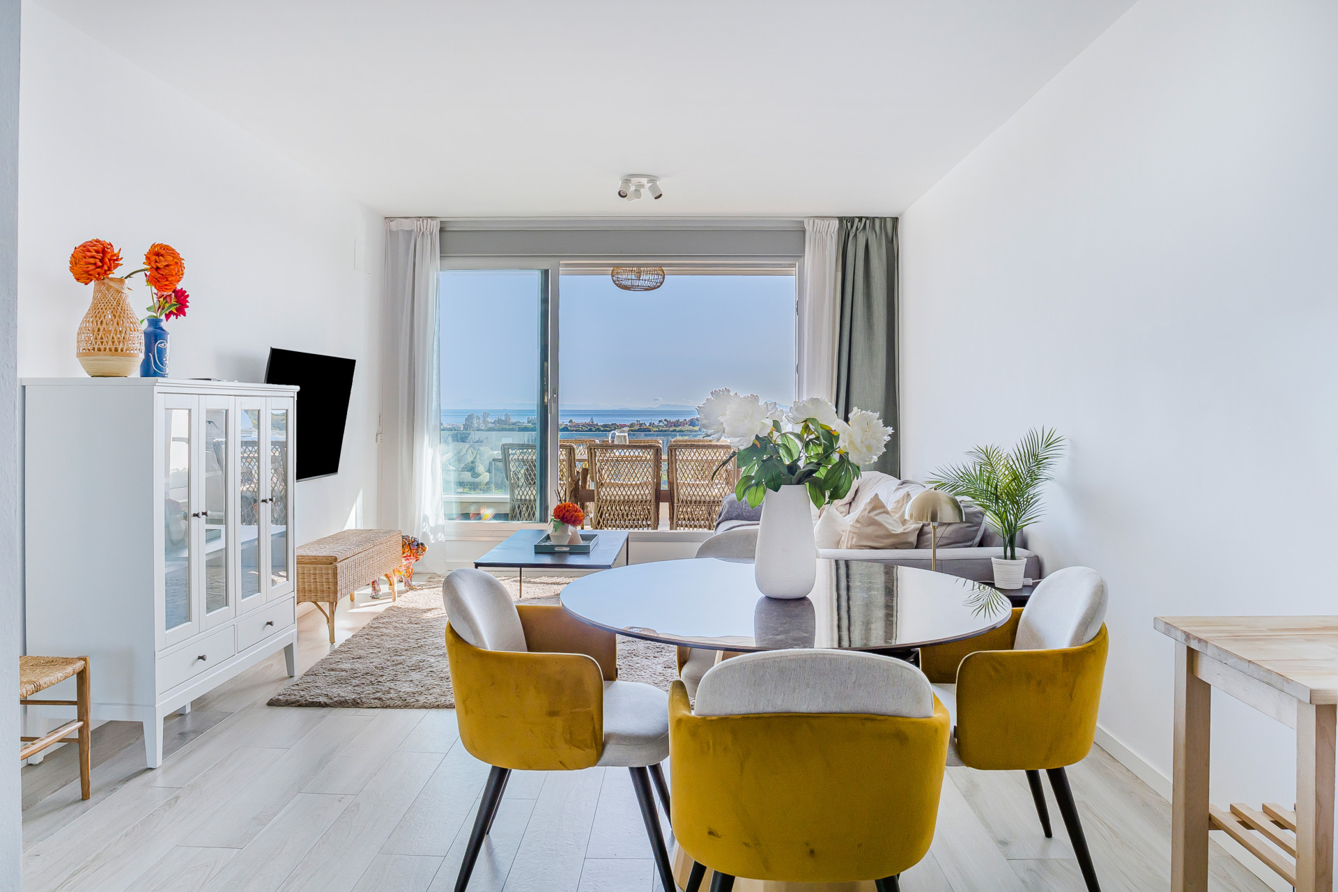 View from dining area to living room with gray sofa and balcony offering ocean and city views.