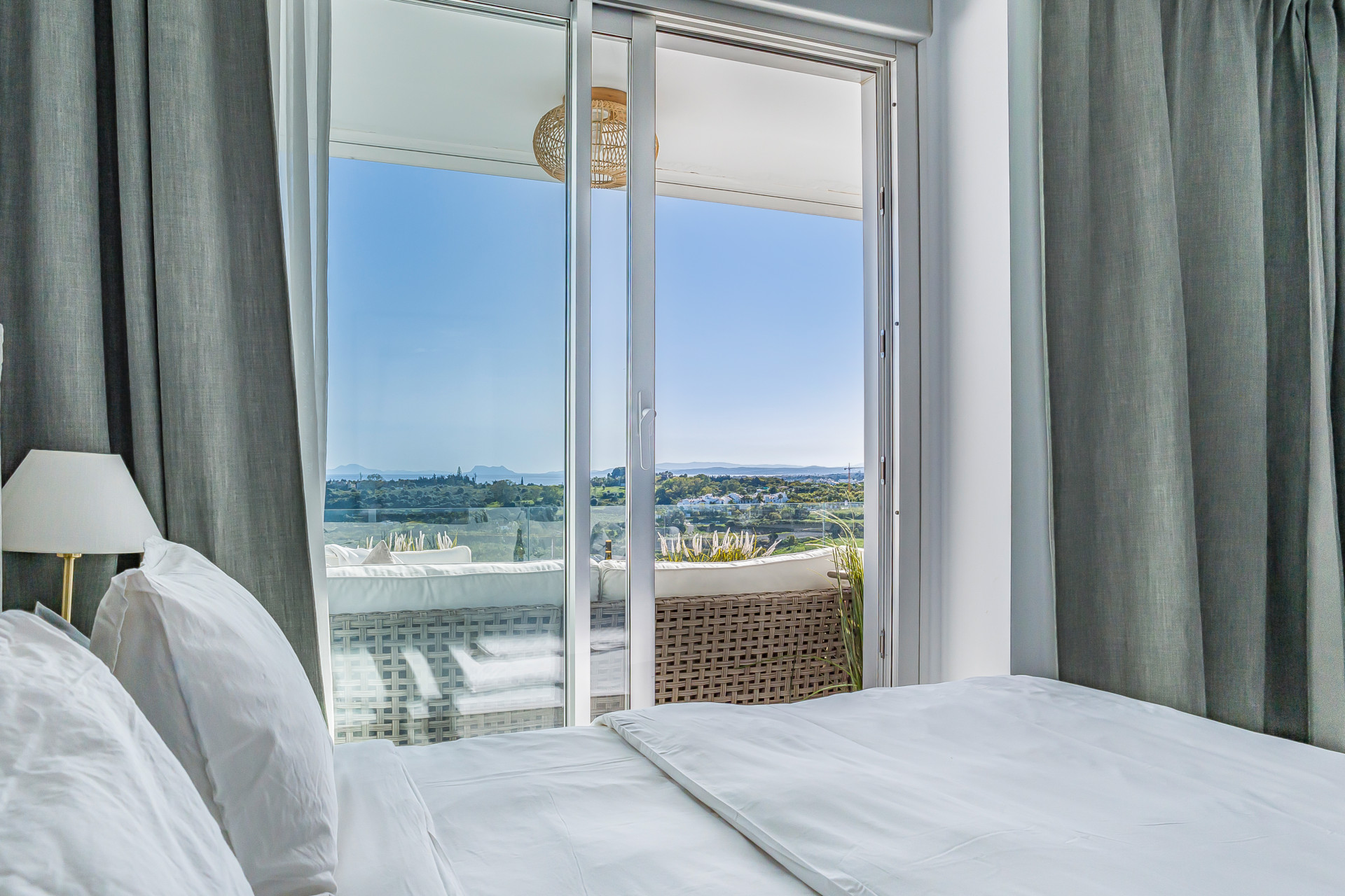 Bedroom with white bedding, gray curtains, and sliding doors opening to a balcony with distant views.