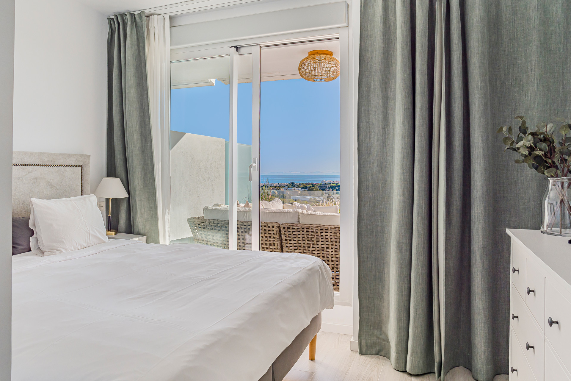 Bedroom with a bed, white dresser, and glass door leading to a balcony with sea views.