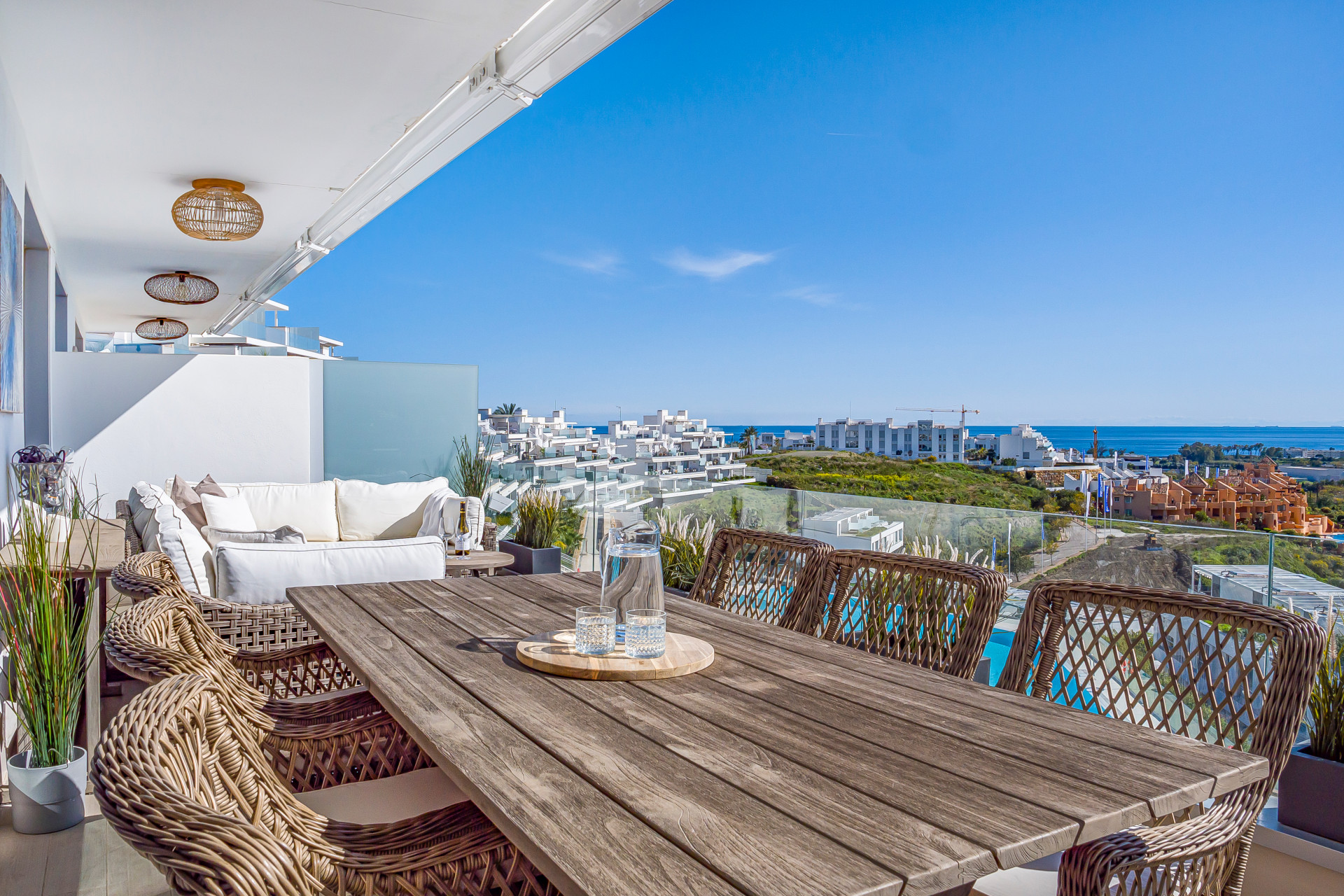Long outdoor dining table with wicker chairs, leading to a white sectional sofa, overlooking the ocean.