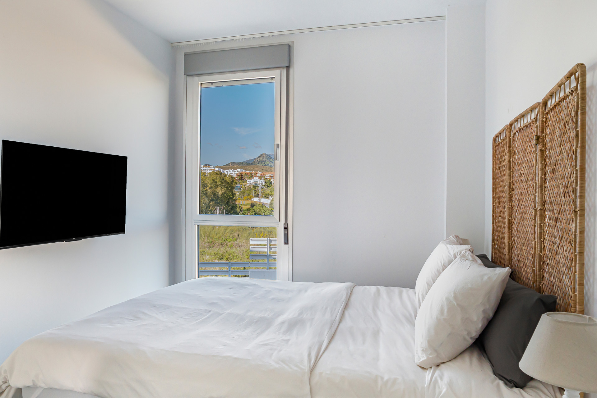 Bedroom with white bedding, wicker headboard, mounted TV, and window view of a town.
