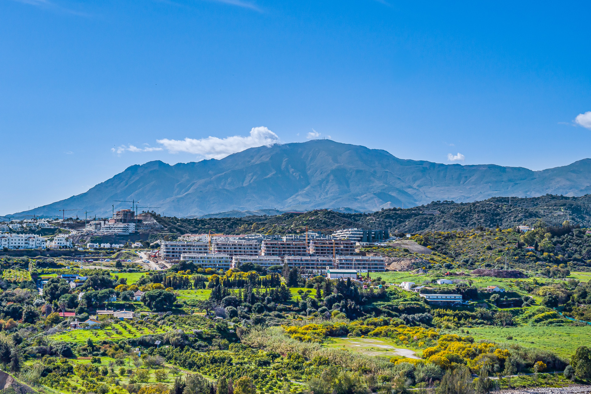 A distant mountain range with construction sites and green landscape in the foreground under a blue sky.