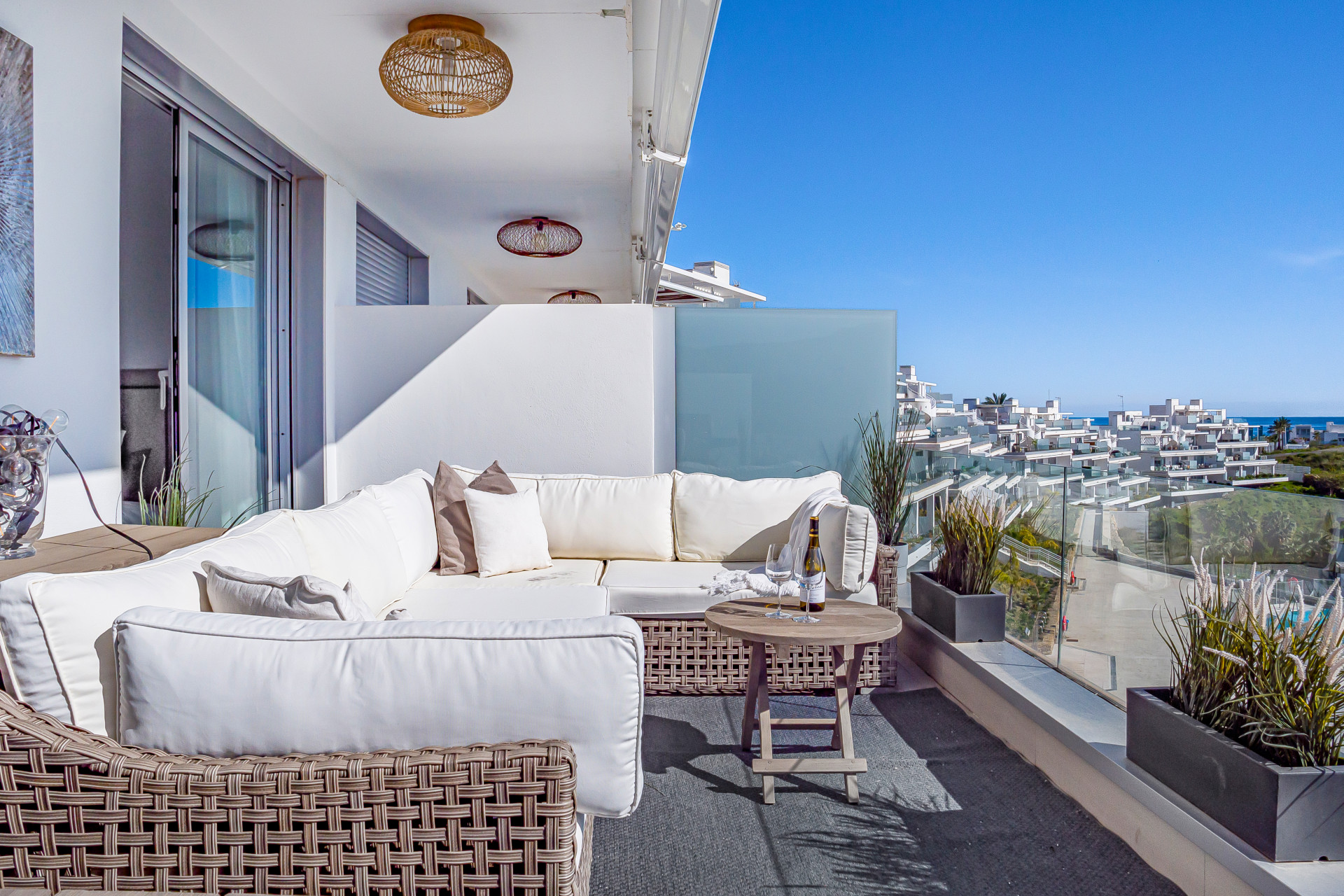 Outdoor balcony with white L-shaped sofa, coffee table, and distant coastal buildings under a bright sky.