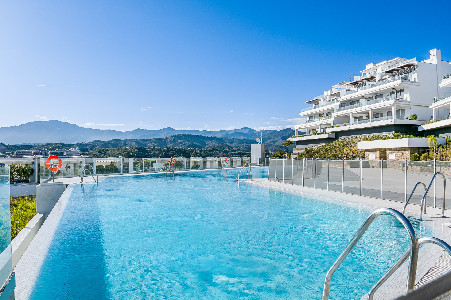 Infinity pool with mountain views, next to a multi-story white apartment building under a clear blue sky.