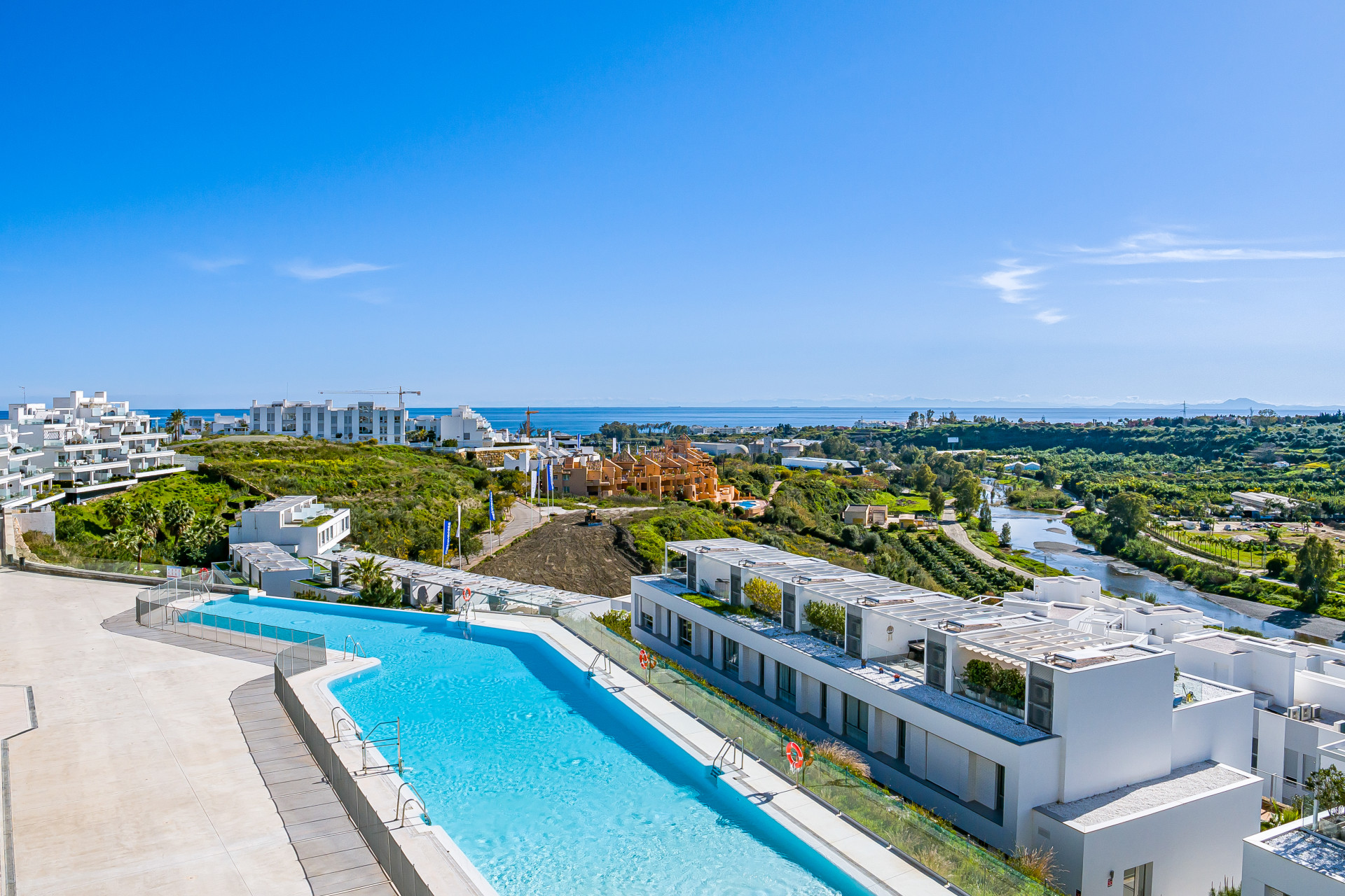 Aerial view of a long infinity pool and white buildings, with ocean and land in the distance.