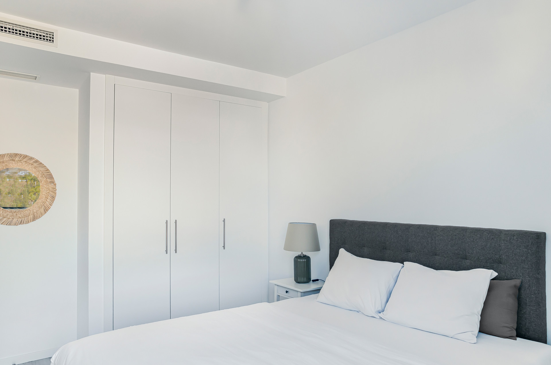 Bedroom view showing white built-in wardrobes, a bed, and a decorative round mirror on the adjacent wall.