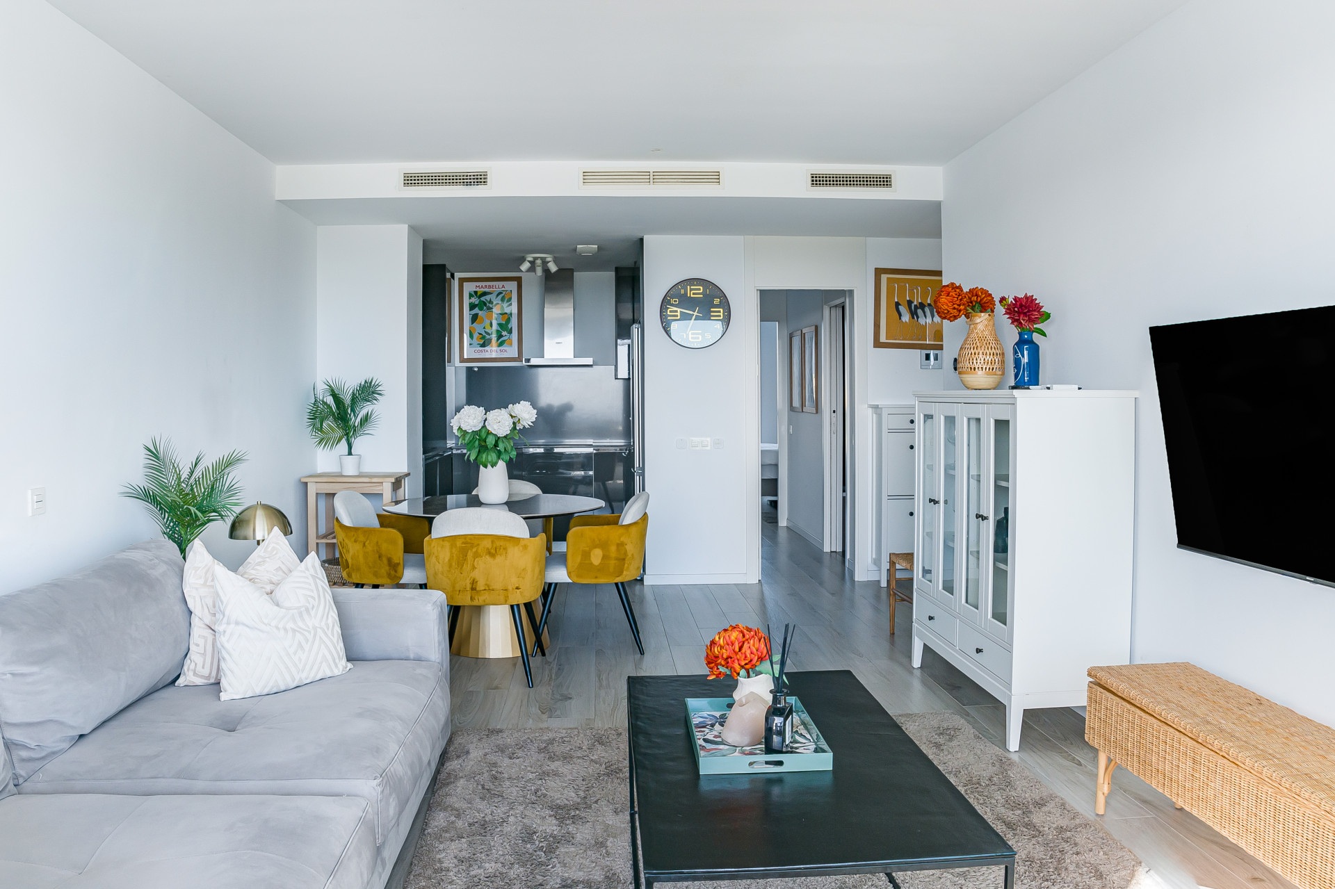 Open plan living area with gray sofa, dining table with yellow chairs, and kitchen in the background.