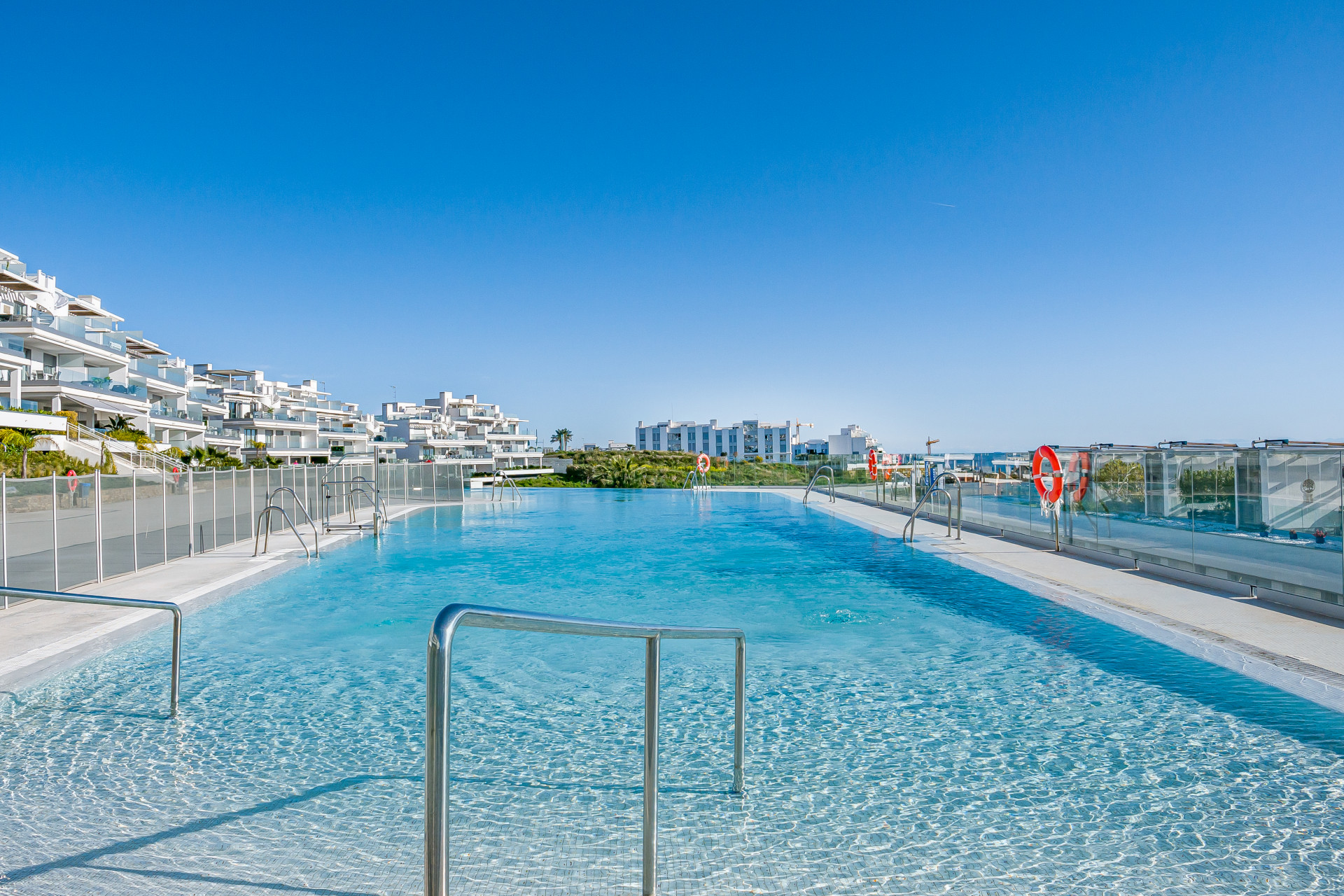 Infinity pool with clear blue water facing modern white buildings under a bright blue sky.