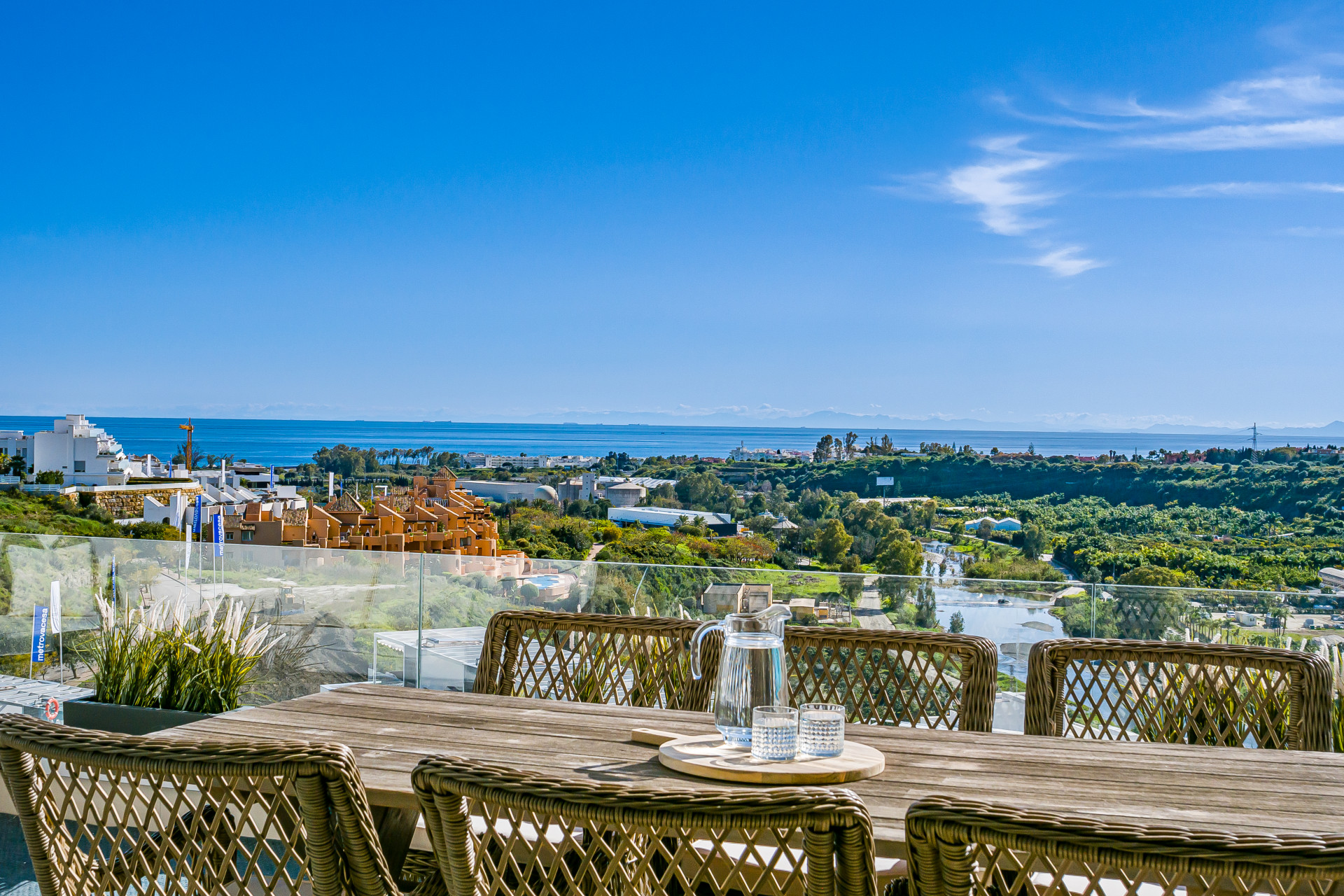 Outdoor dining table with wicker chairs, pitcher, glasses, and a wide view of the sea and coastline.