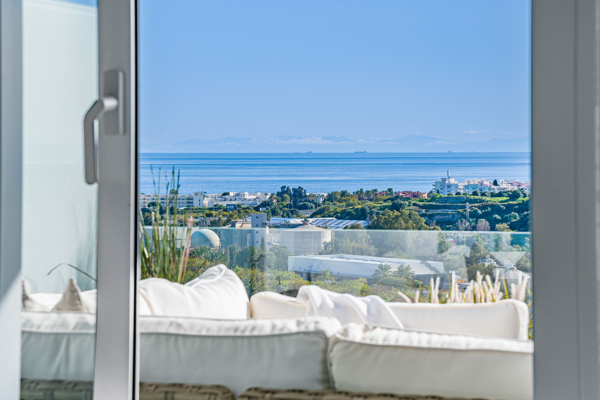 Balcony view over rooftops to the sea and distant mountains under a blue sky.