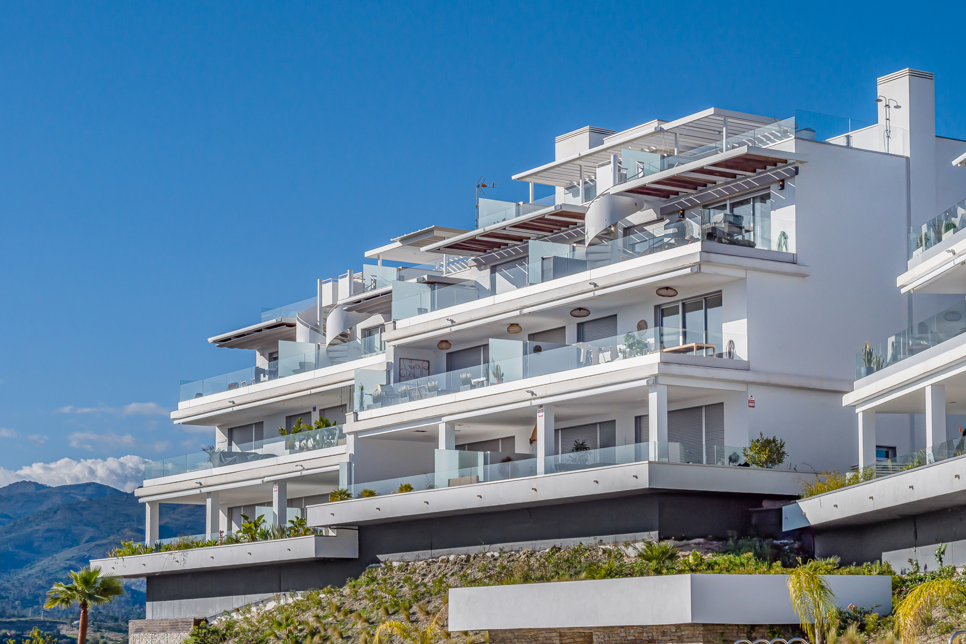 Exterior view of a modern tiered apartment building with glass balconies, built on a sloped landscape.