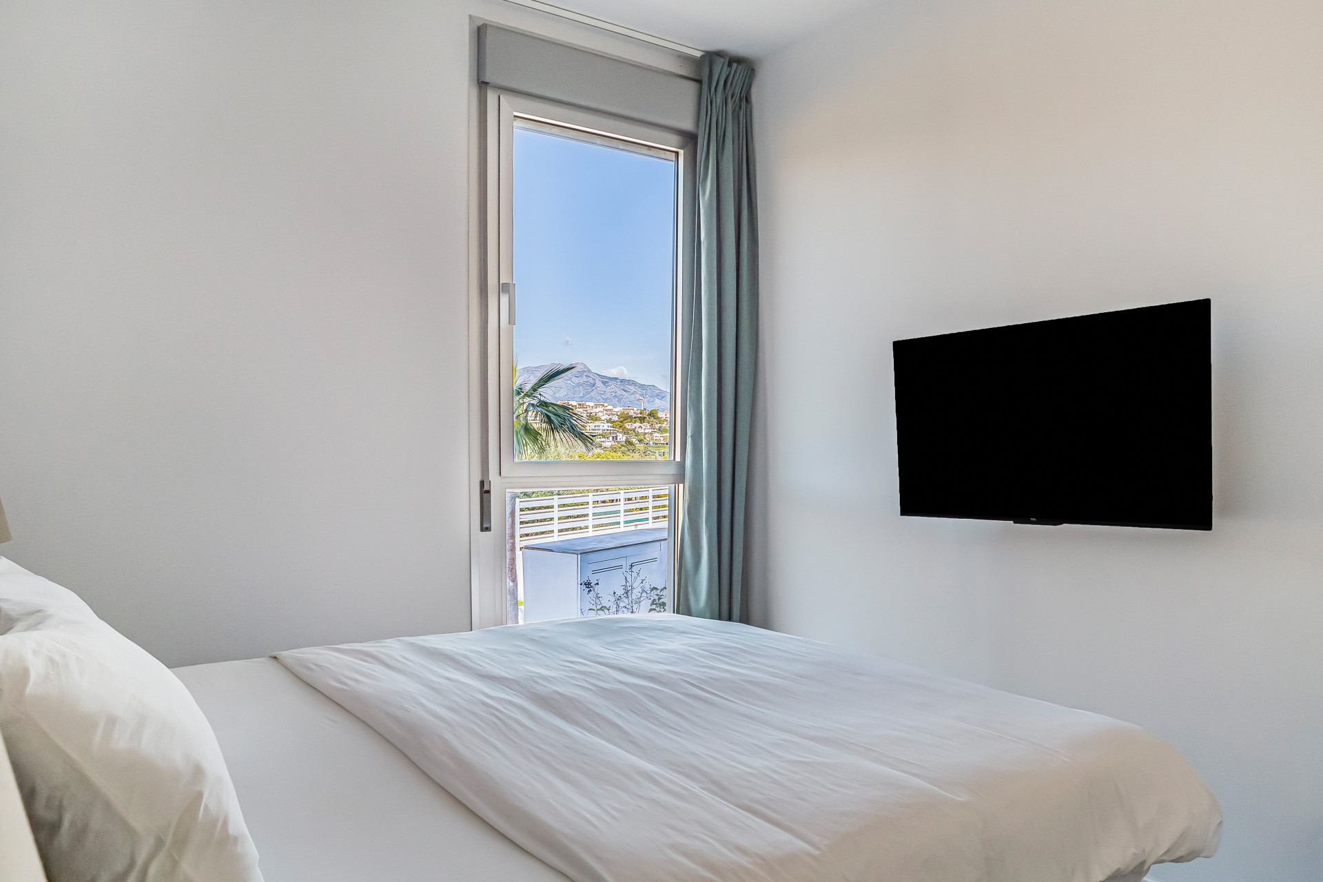 Bedroom featuring a wall-mounted TV, a bed with white bedding, and a full-height window overlooking mountains.