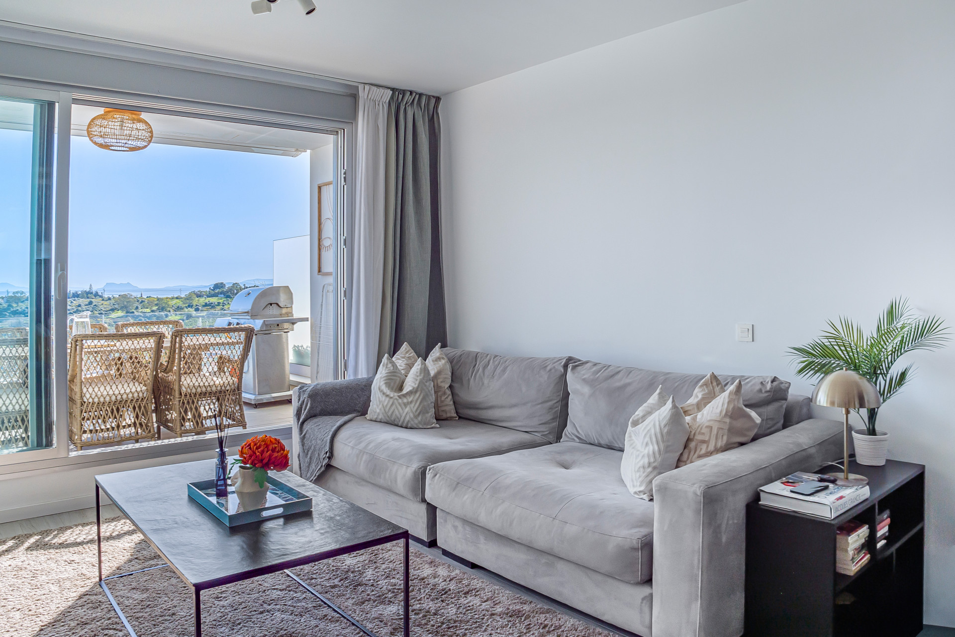 Living room featuring a large gray sofa, coffee table, and an open balcony with outdoor furniture.
