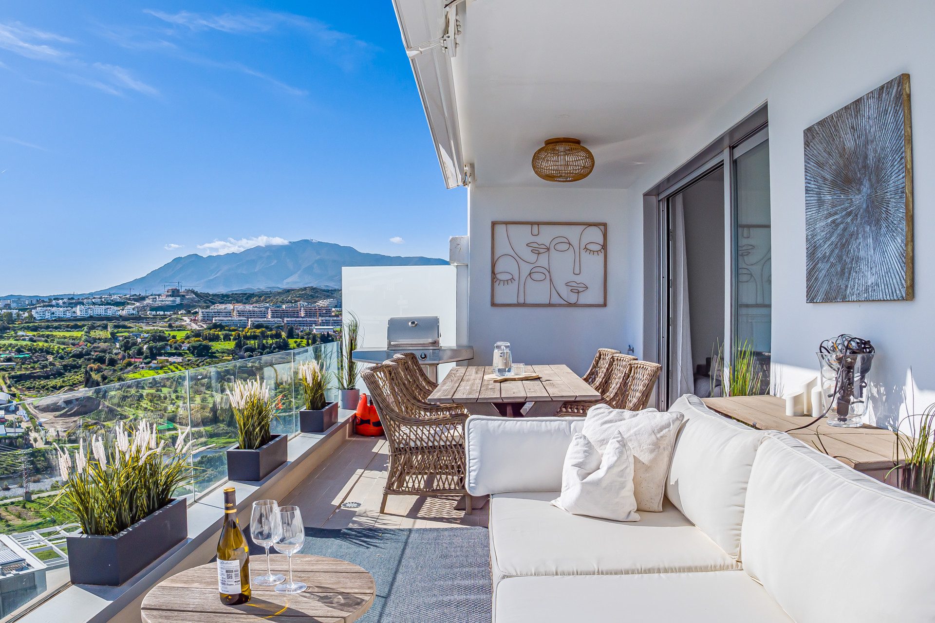 Large outdoor balcony with white sofa, dining table, grill, and panoramic mountain views under a blue sky.