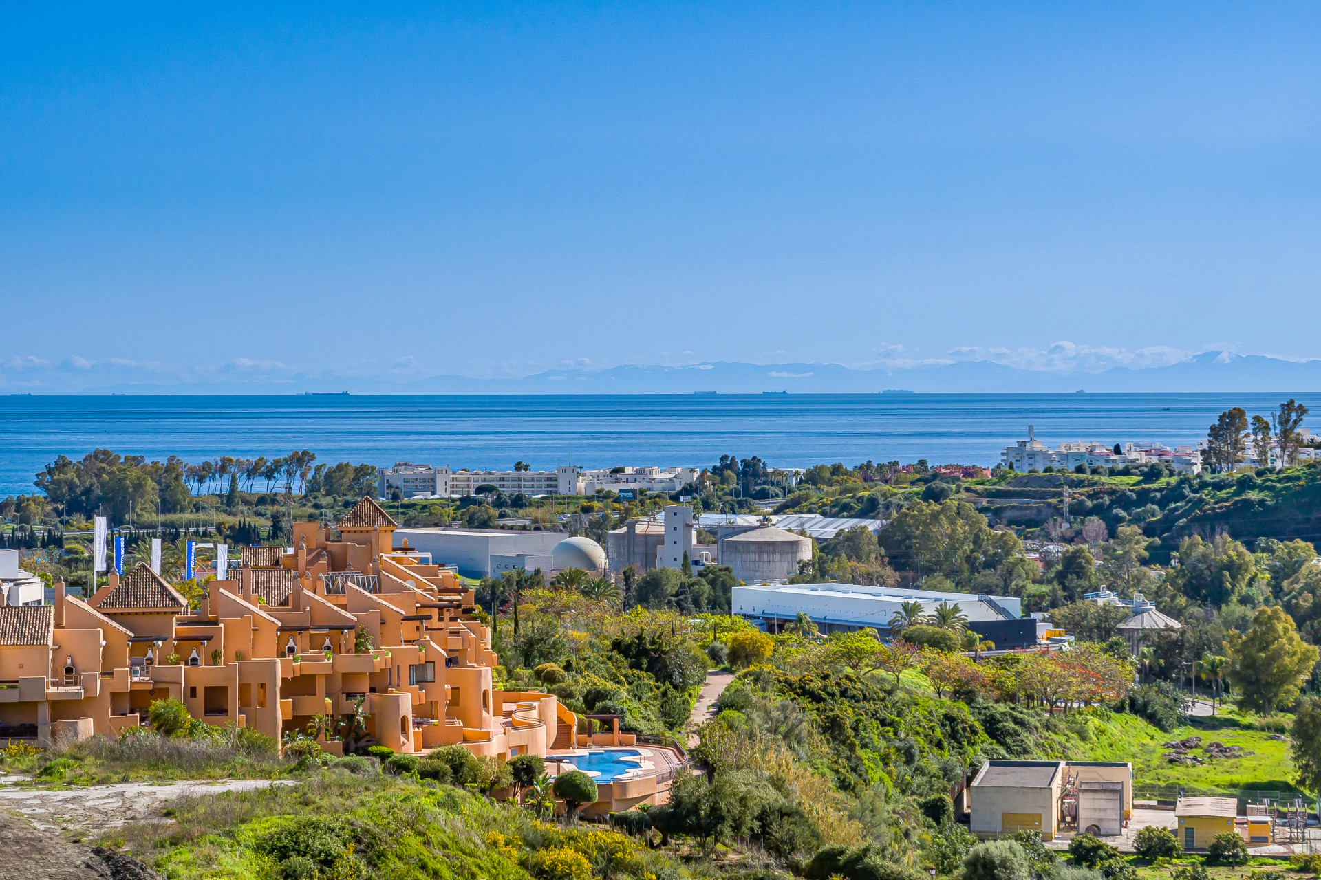 Panoramic view of a coastal town with buildings, green hills, and the Mediterranean Sea under blue sky.