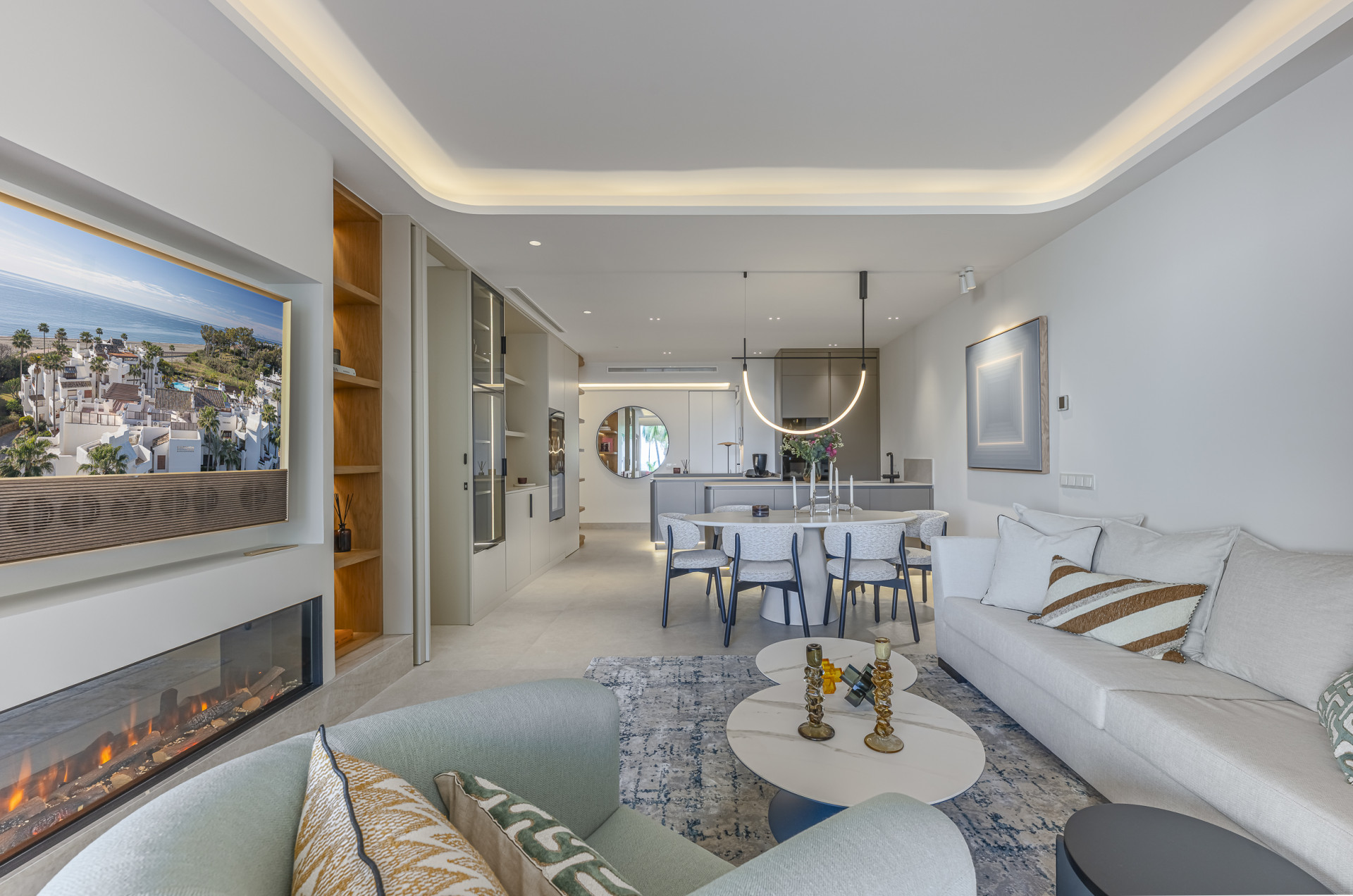 Open plan living area with a white sofa, armchair, coffee tables, and a dining area with kitchen beyond.