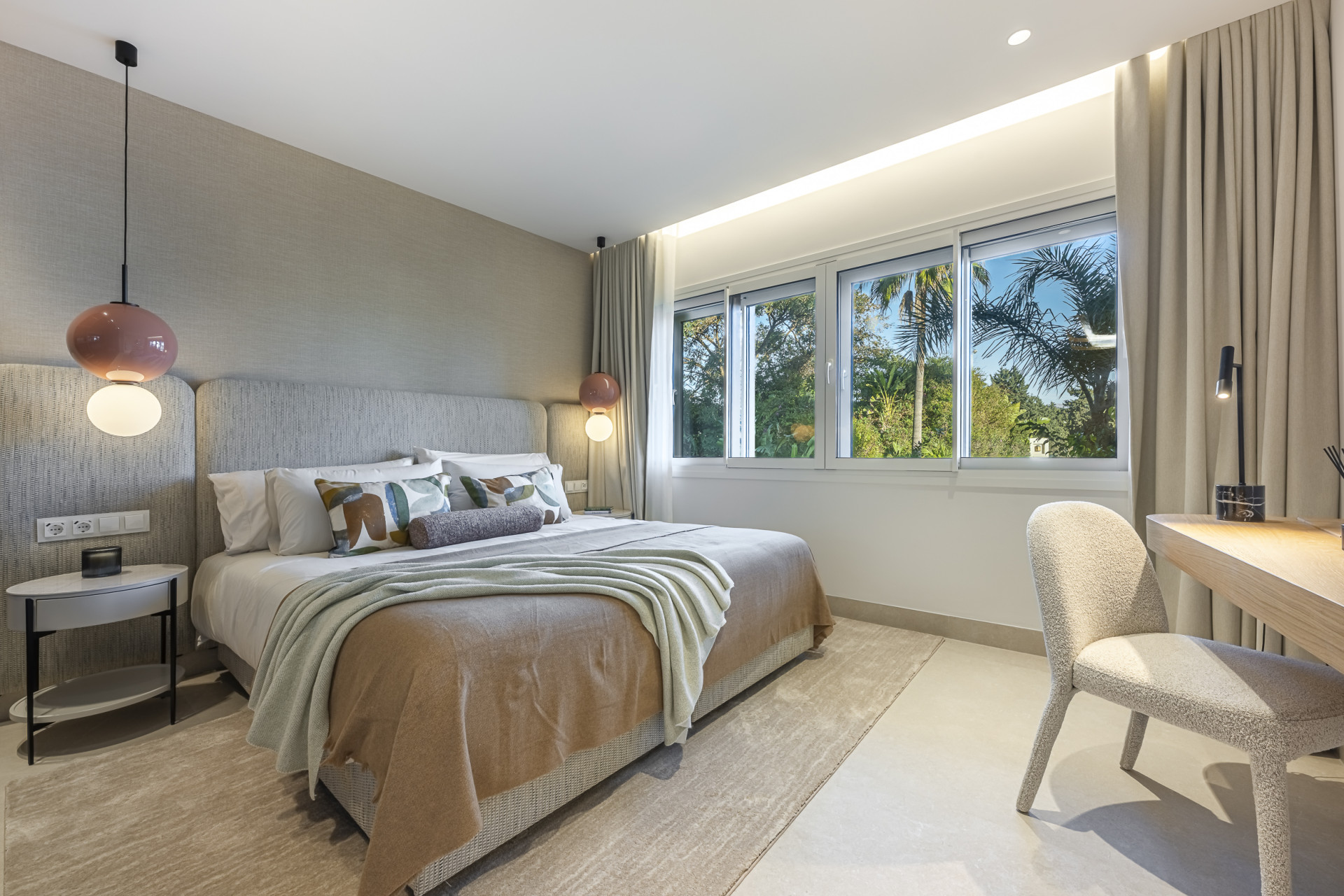 Bedroom featuring a large bed, side tables with lamps, a desk, chair, and large windows with greenery views.