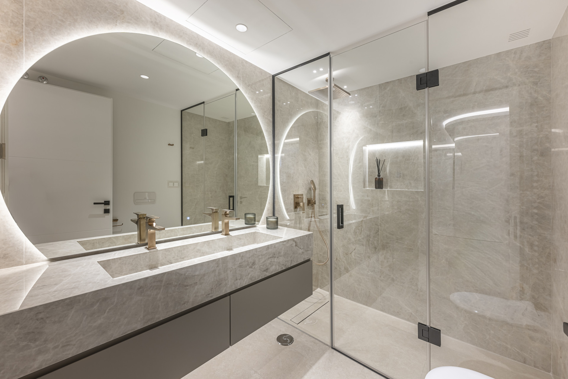 Bathroom with a large round backlit mirror, double vanity, and a glass-enclosed shower.