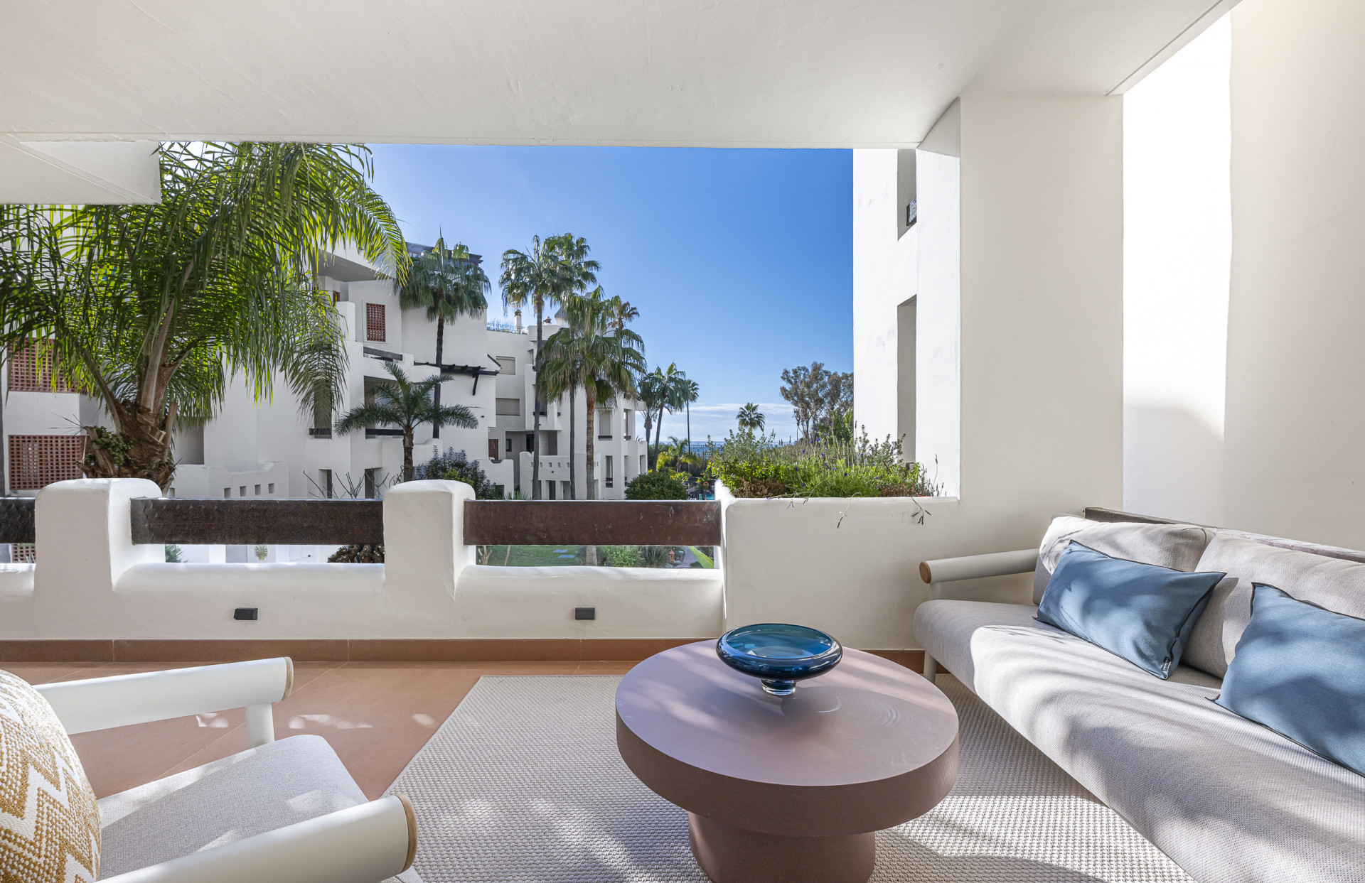 Outdoor patio with light grey sofa, armchair, and coffee table, facing a garden view.