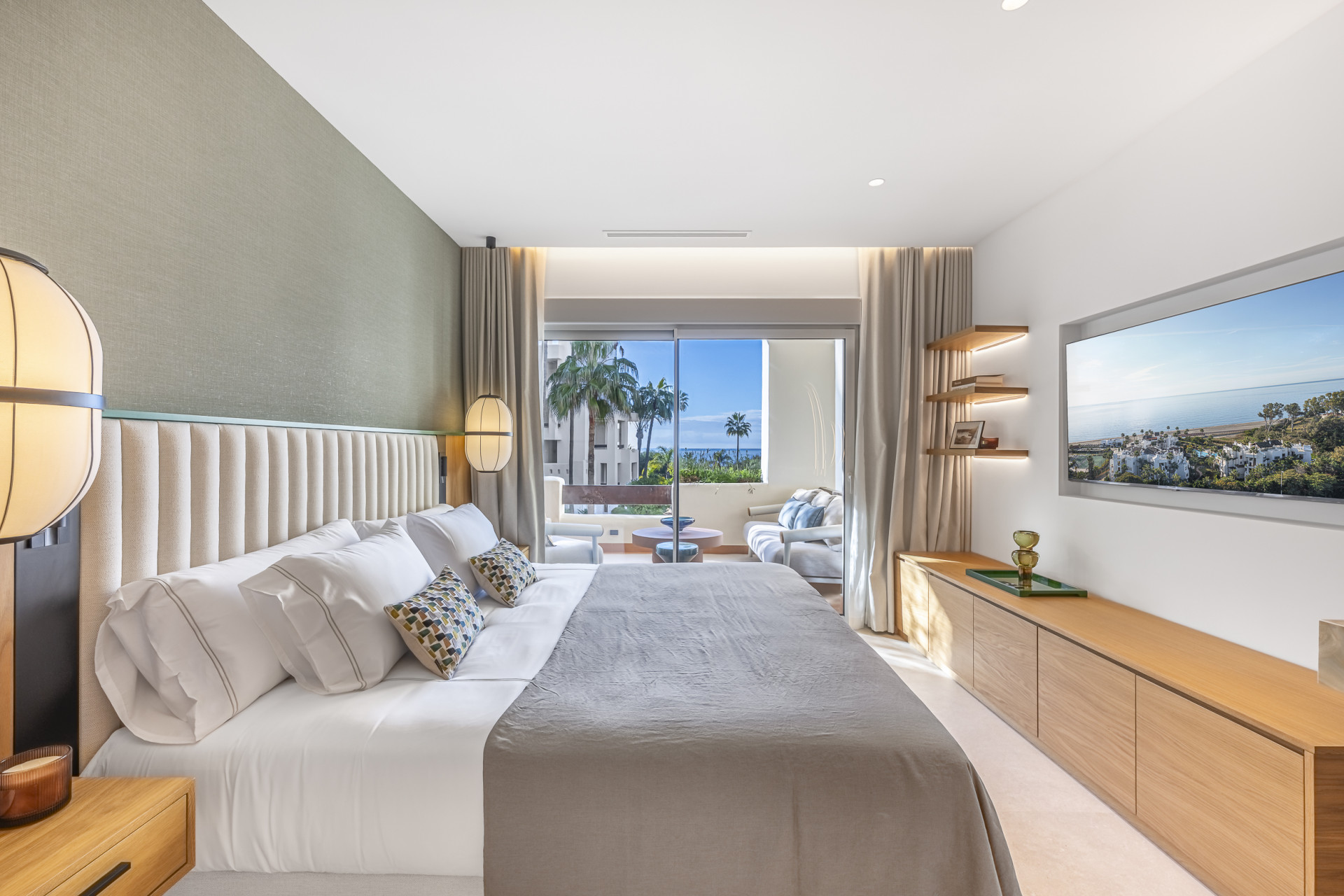 Bedroom with king-size bed, TV, light green accent wall, and a balcony view of palm trees and ocean.