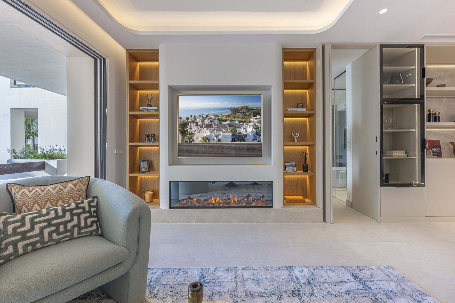 View of a living room wall with a large TV, fireplace, built-in wooden shelves, and an open glass cabinet.