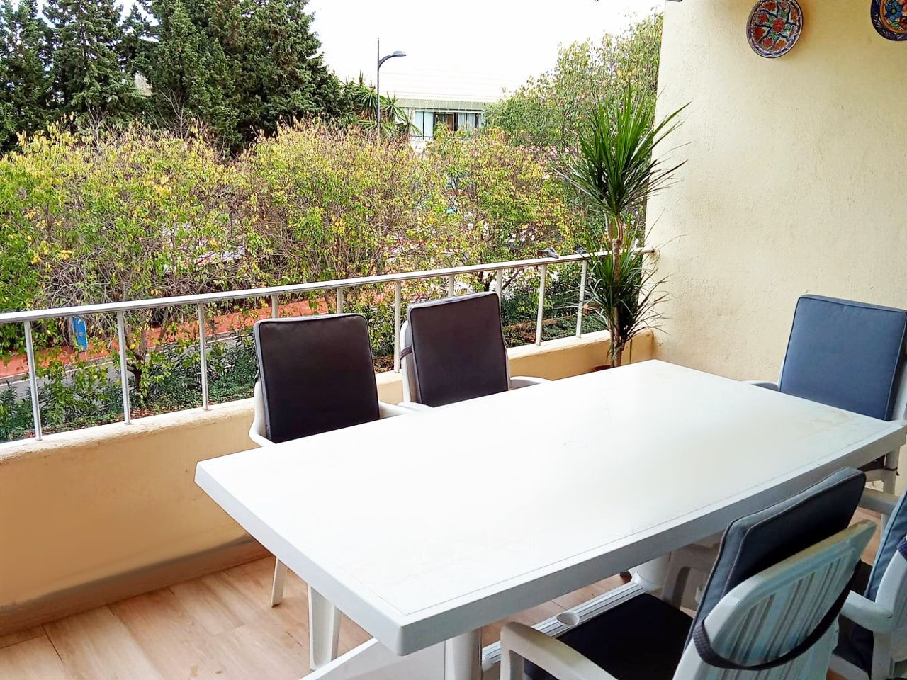 Outdoor balcony with a white dining table, four chairs, and lush green trees visible beyond the railing.