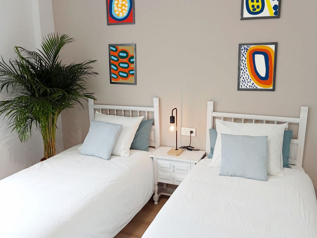 Close up of two single beds with white linens, light blue pillows, and colorful framed art on a beige wall.