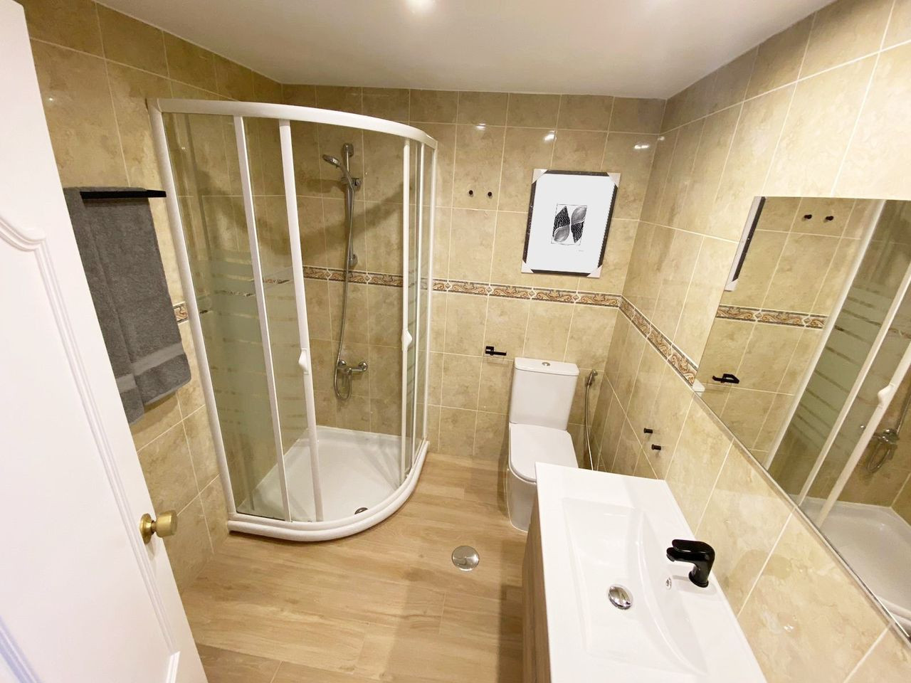 Bathroom with tan tiled walls and floor, corner shower, toilet, white vanity with sink, and mirror.