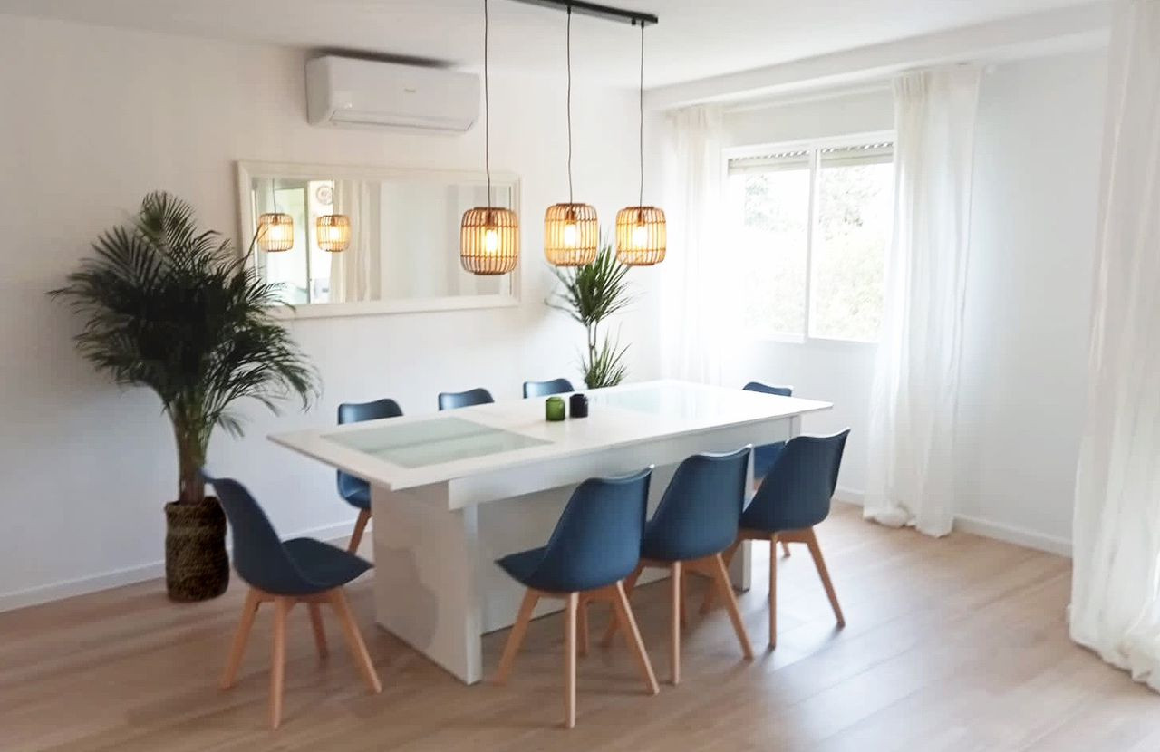 Dining area with a long white table, eight blue chairs, and three rattan pendant lights.