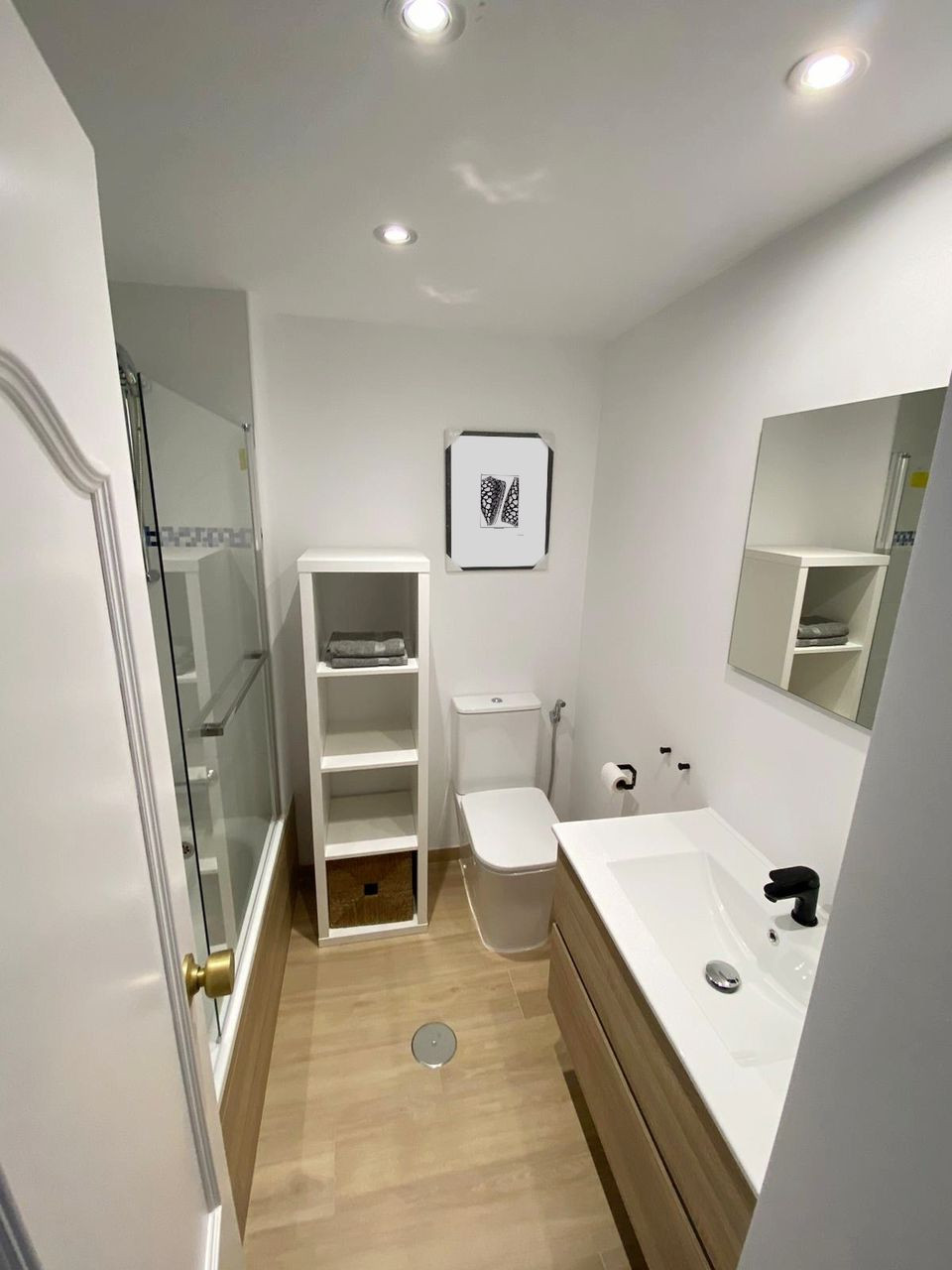 Bathroom with light wood floor, white vanity with black faucet, toilet, shower tub, and white shelving unit.