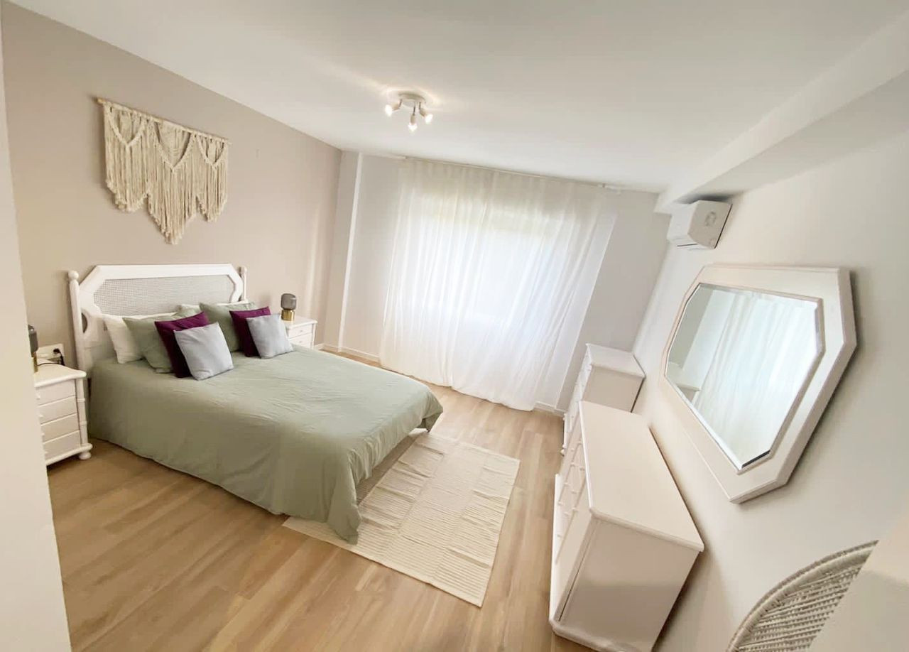 Bedroom with light wood floor, light green bed, white nightstands, dressers, mirror, and sheer curtains.