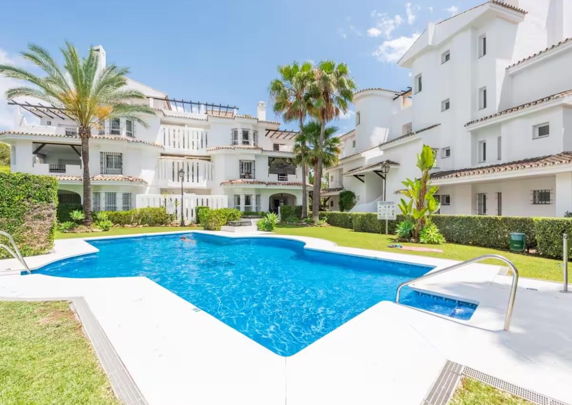 Outdoor pool surrounded by white buildings, green lawn, and palm trees under a clear sky.