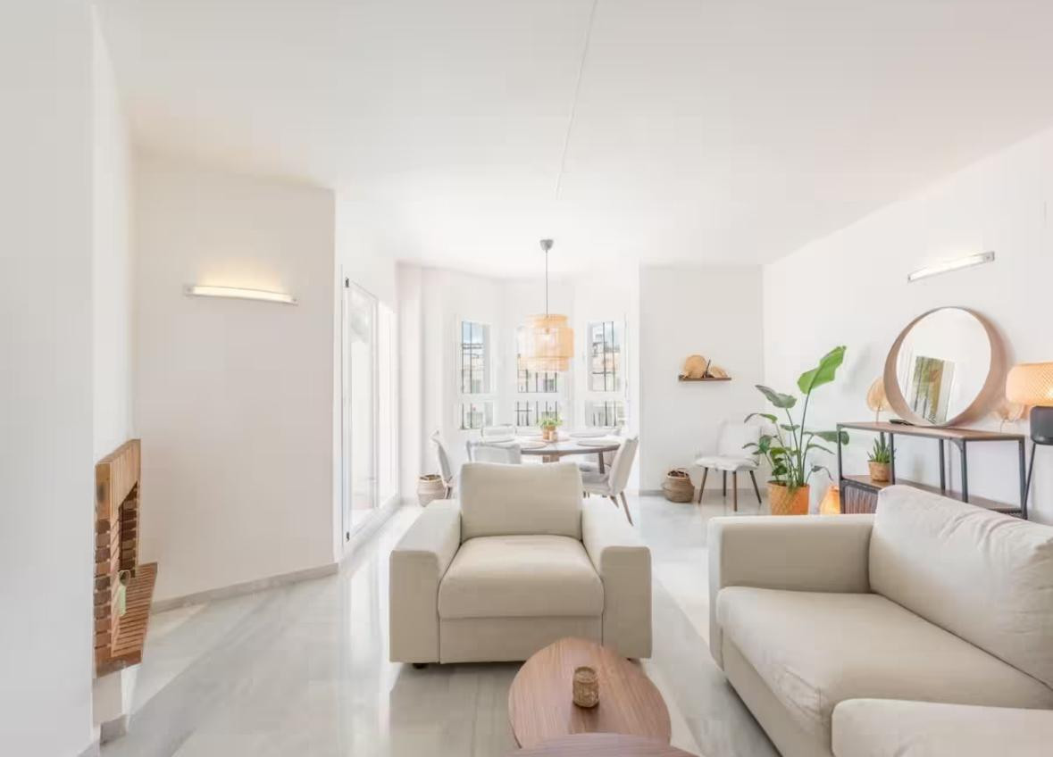 Bright living room with light sofas, coffee table, and a dining area in the background with a circular table.