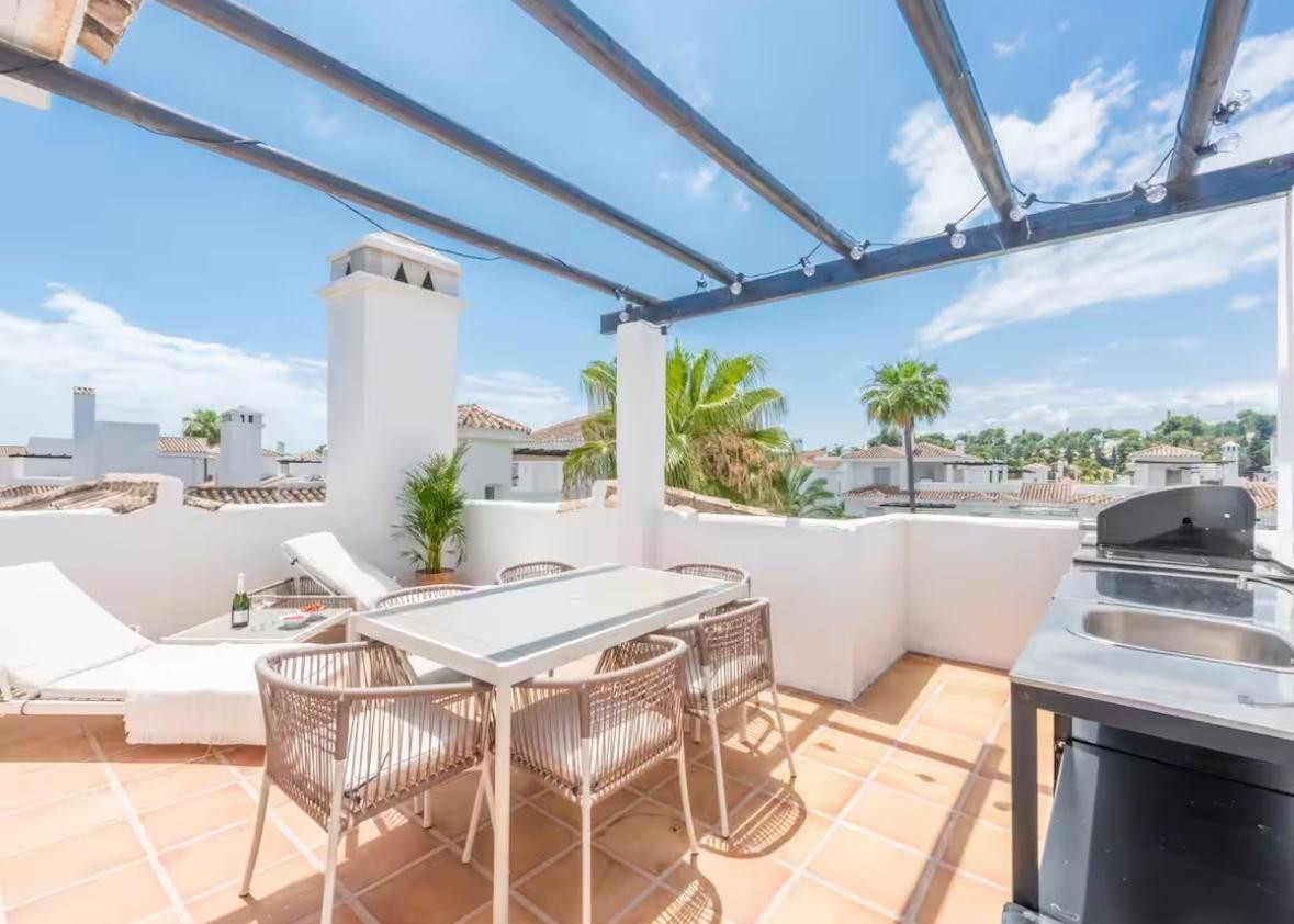 Rooftop terrace with an outdoor dining table, grill, and lounge chairs under a pergola.