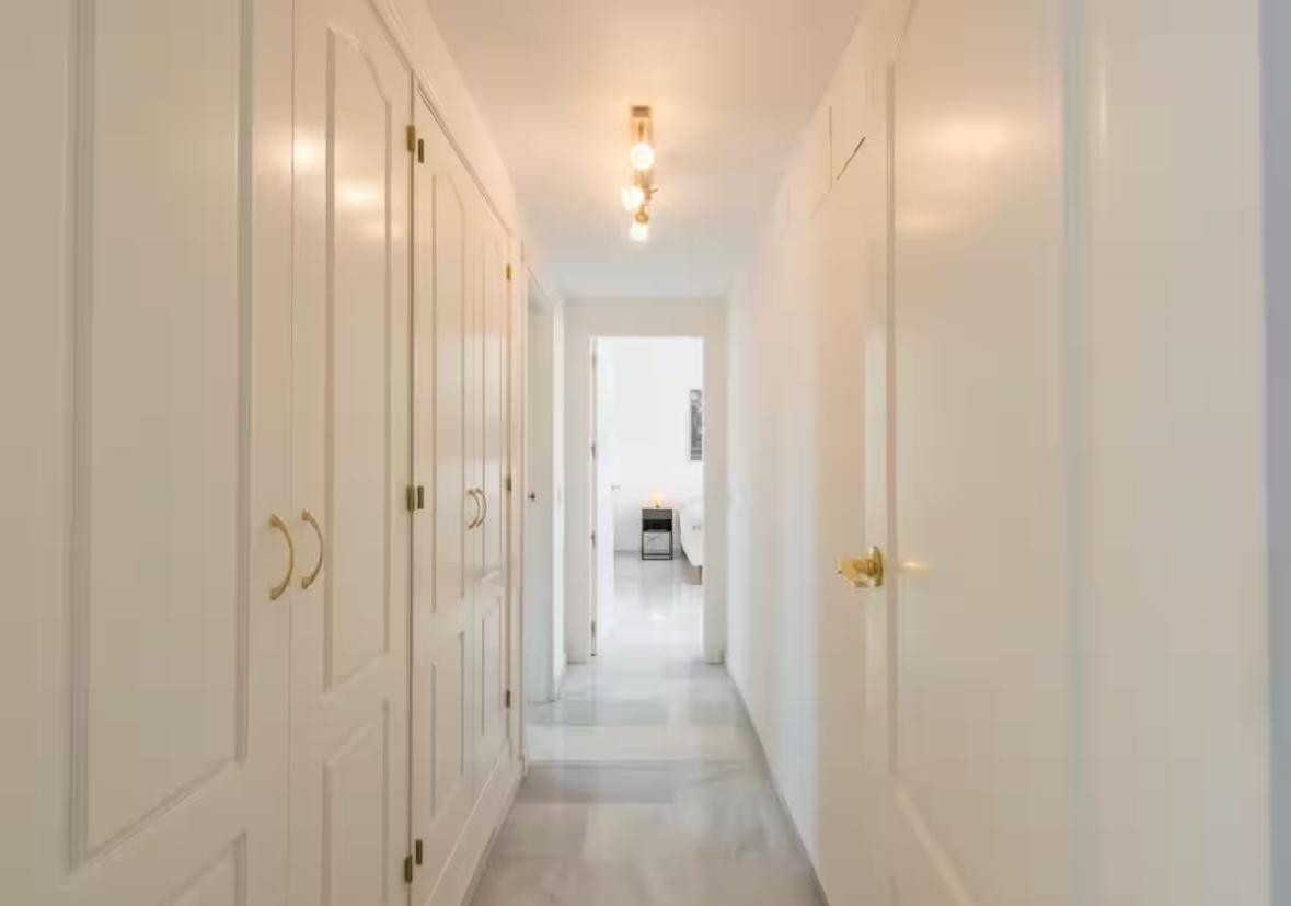 Narrow hallway with white built-in closets on the left and a series of ceiling lights leading to another room.
